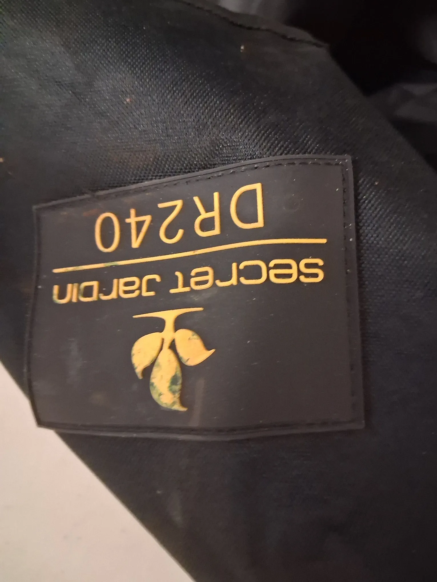 Zbliżenie na czarną etykietę z złotymi napisami i grafiką, przymocowaną do ciemnego materiału. Na etykiecie znajduje się pionowy napis „secret jardin” oraz oznaczenie „DR240”. Po lewej stronie widnieje złoty symbol przypominający gałązkę z liśćmi.