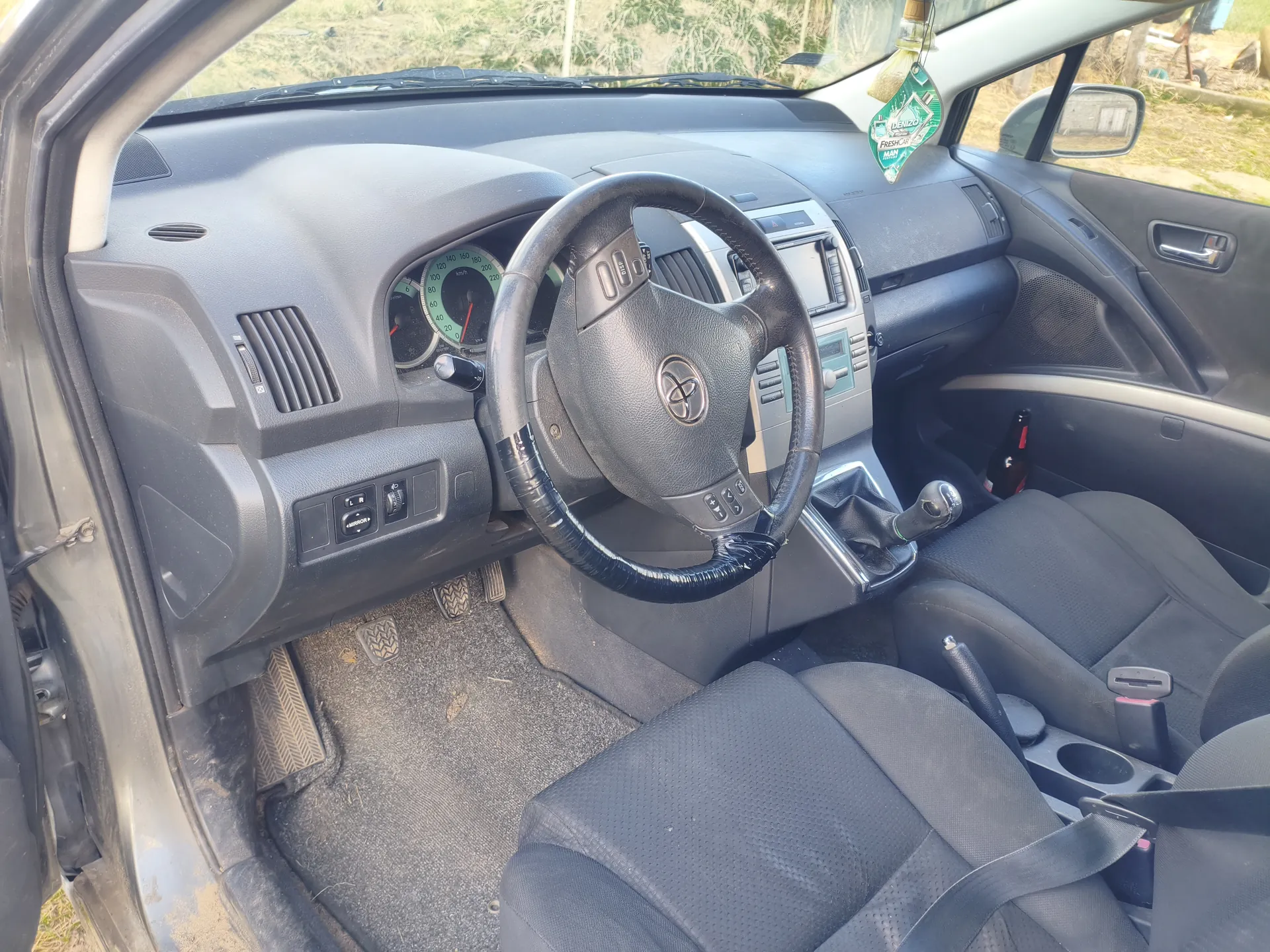 Wnętrze samochodu Toyota z widoczną kierownicą z logo marki, tablicą rozdzielczą z prędkościomierzem do 220 km/h, czarnymi siedzeniami, dźwignią zmiany biegów i zielonym odświeżaczem powietrza wiszącym na lusterku wstecznym. Drzwi kierowcy są otwarte, a na podłodze widoczne są brudne dywaniki. W kieszeni drzwi znajduje się butelka.