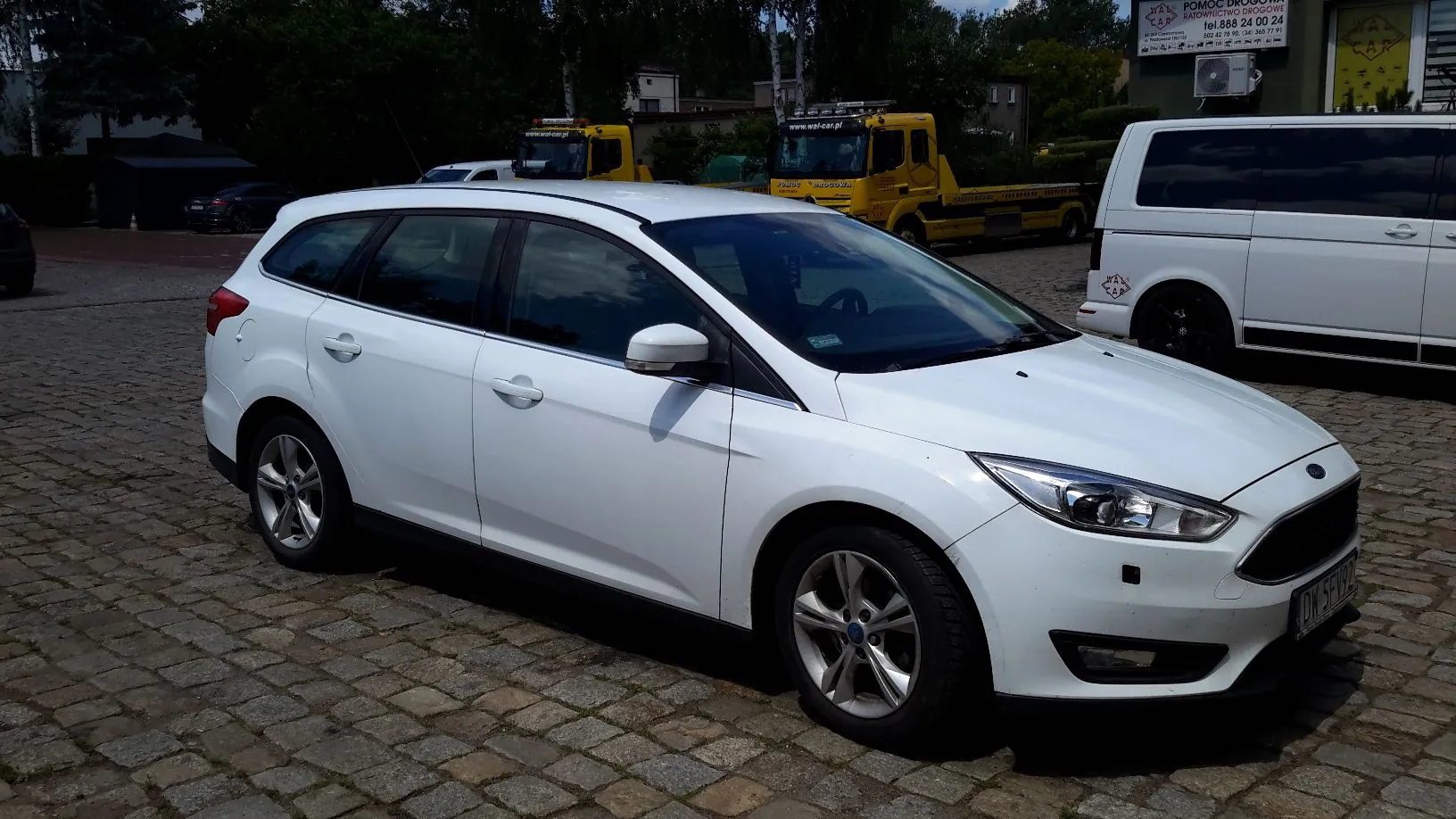 Biały samochód osobowy typu kombi marki Ford zaparkowany na kostce brukowej. W tle widoczne są żółte samochody ciężarowe z napisami 'www.wsj-car.pl' oraz biały samochód dostawczy. W oddali znajdują się budynki i drzewa.