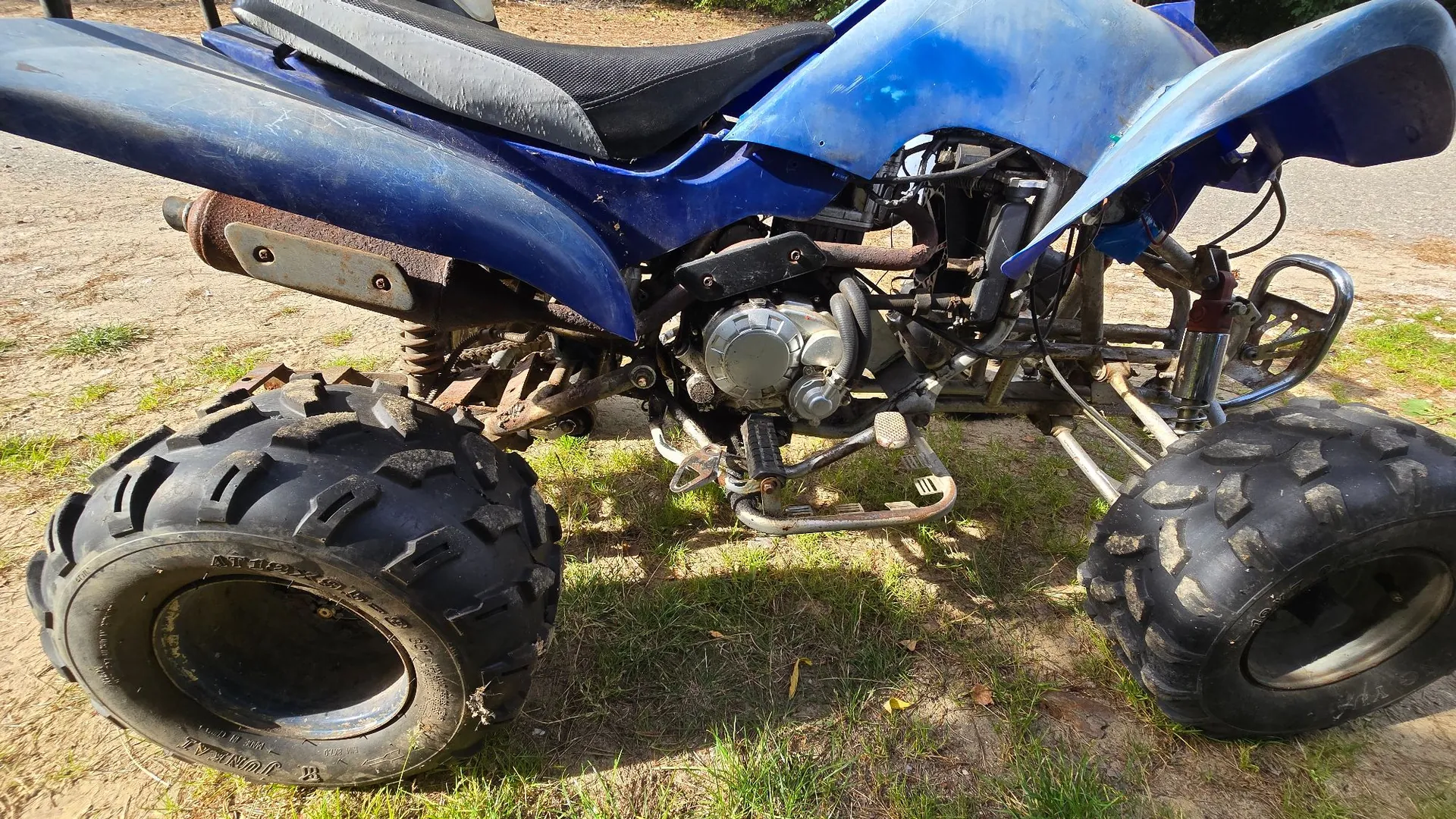 Niebieski quad terenowy zaparkowany na trawiasto-piaszczystym podłożu. Widoczny jest z boku, ukazując szerokie, terenowe opony z głębokim bieżnikiem, silnik, rurę wydechową oraz czarne siedzenie.