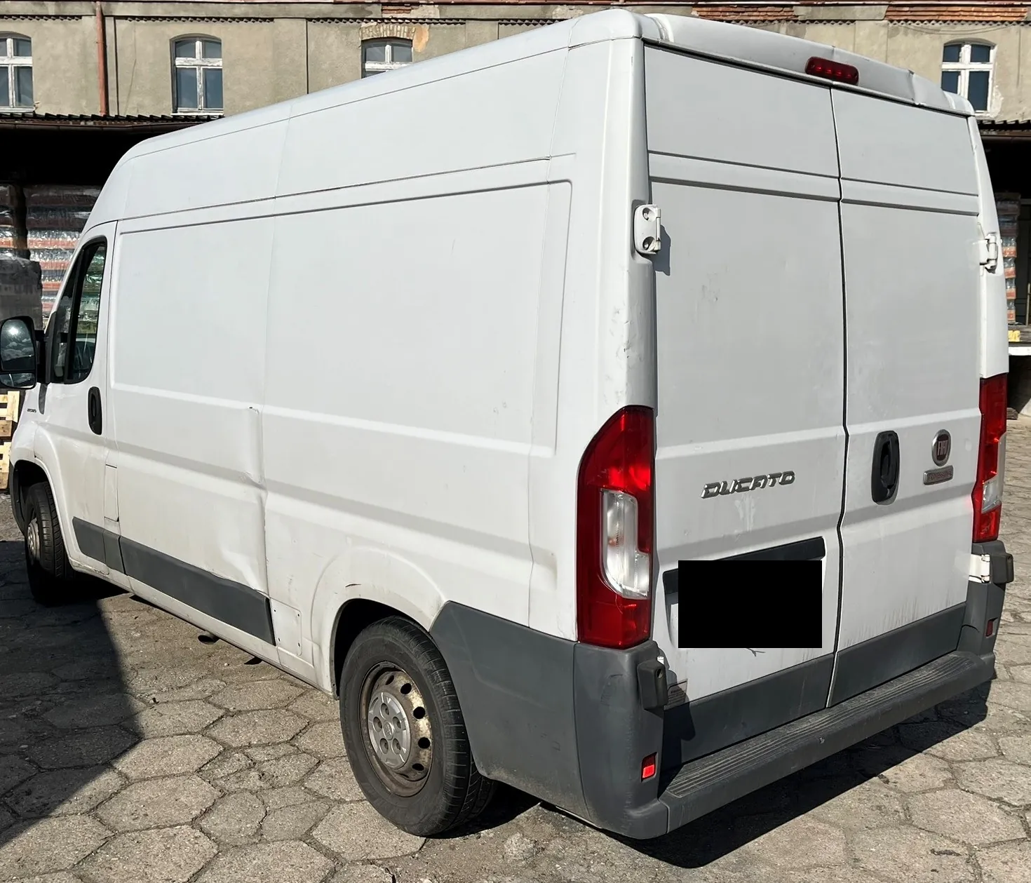 Biały furgon marki Fiat Ducato zaparkowany na kostce brukowej na tle starego budynku przemysłowego. Pojazd jest widoczny od tyłu i z lewej strony. Na tylnych drzwiach widnieje napis 'DUCATO' oraz logo Fiata. Na lewym boku nadwozia widoczne jest wgniecenie. Tablica rejestracyjna jest zakryta czarnym prostokątem.