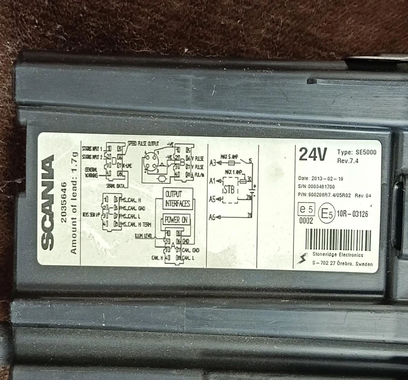 Czarny przedmiot z białą etykietą. Na etykiecie widnieje napis 'SCANIA', numer 2035646, informacja 'Amount of lead: 1.7g', schematy elektryczne, oznaczenia '24V', 'Type: SE5000', 'Rev 7.4', numery seryjne i kreskowy kod kreskowy. W dolnej części etykiety znajduje się napis 'Stoneridge Electronics' oraz adres 'S-702 27 Örebro, Sweden'.