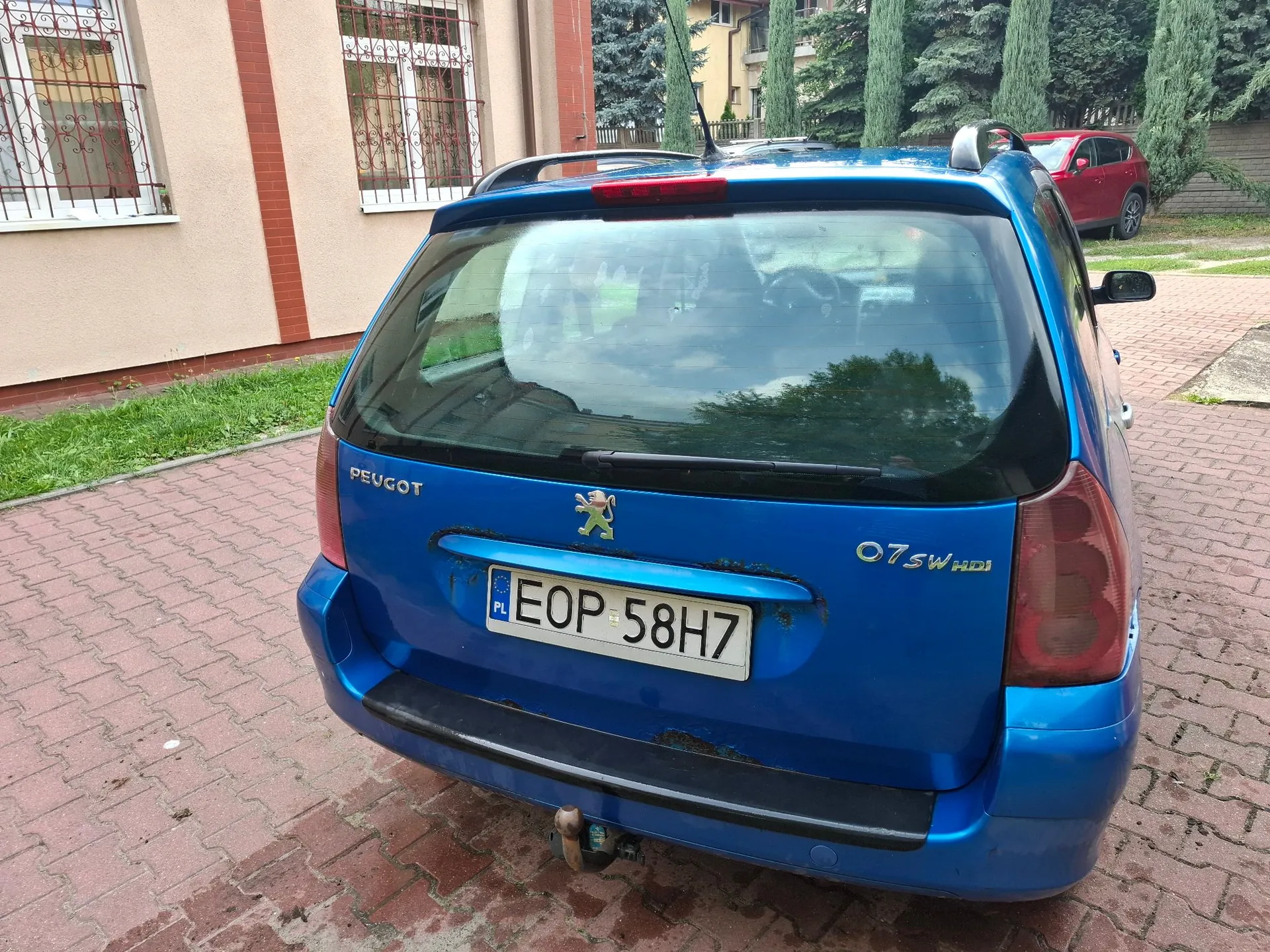 Tylny widok niebieskiego samochodu marki Peugeot, model 206 SW HDI. Pojazd posiada polską tablicę rejestracyjną o numerze EOP 58H7. Samochód zaparkowany jest na kostce brukowej obok budynku o beżowej elewacji z oknami zabezpieczonymi kratami. W tle widoczne są zielone drzewa i czerwony samochód.