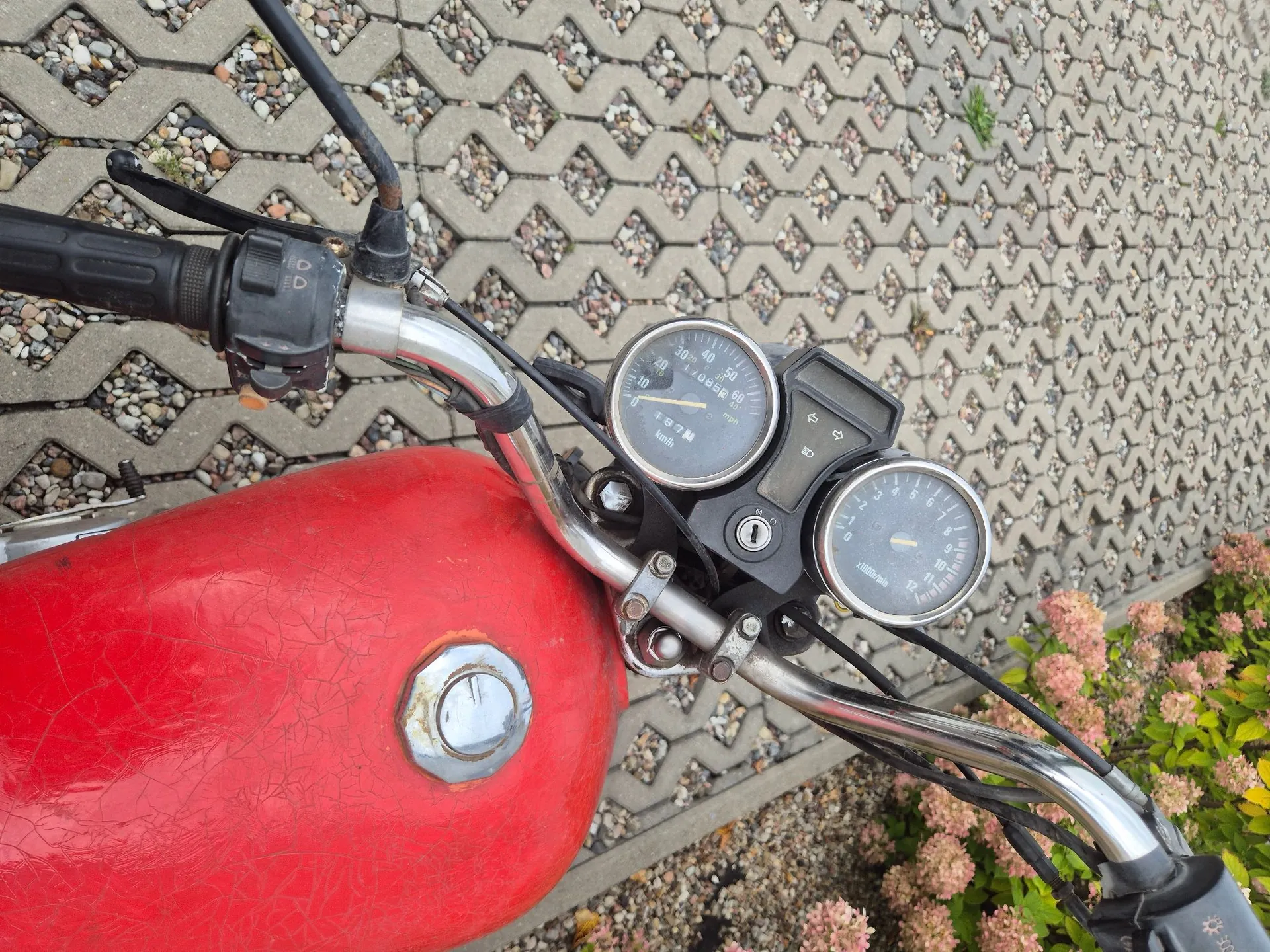 Widok z perspektywy kierowcy na kierownicę i czerwony bak motocykla. Na kierownicy znajdują się dwa analogowe wskaźniki: prędkościomierz z podziałką do 60 km/h oraz obrotomierz. Widoczny jest metalowy uchwyt kierownicy, manetka oraz stacyjka. Motocykl stoi na kostce brukowej z trawą w szczelinach.