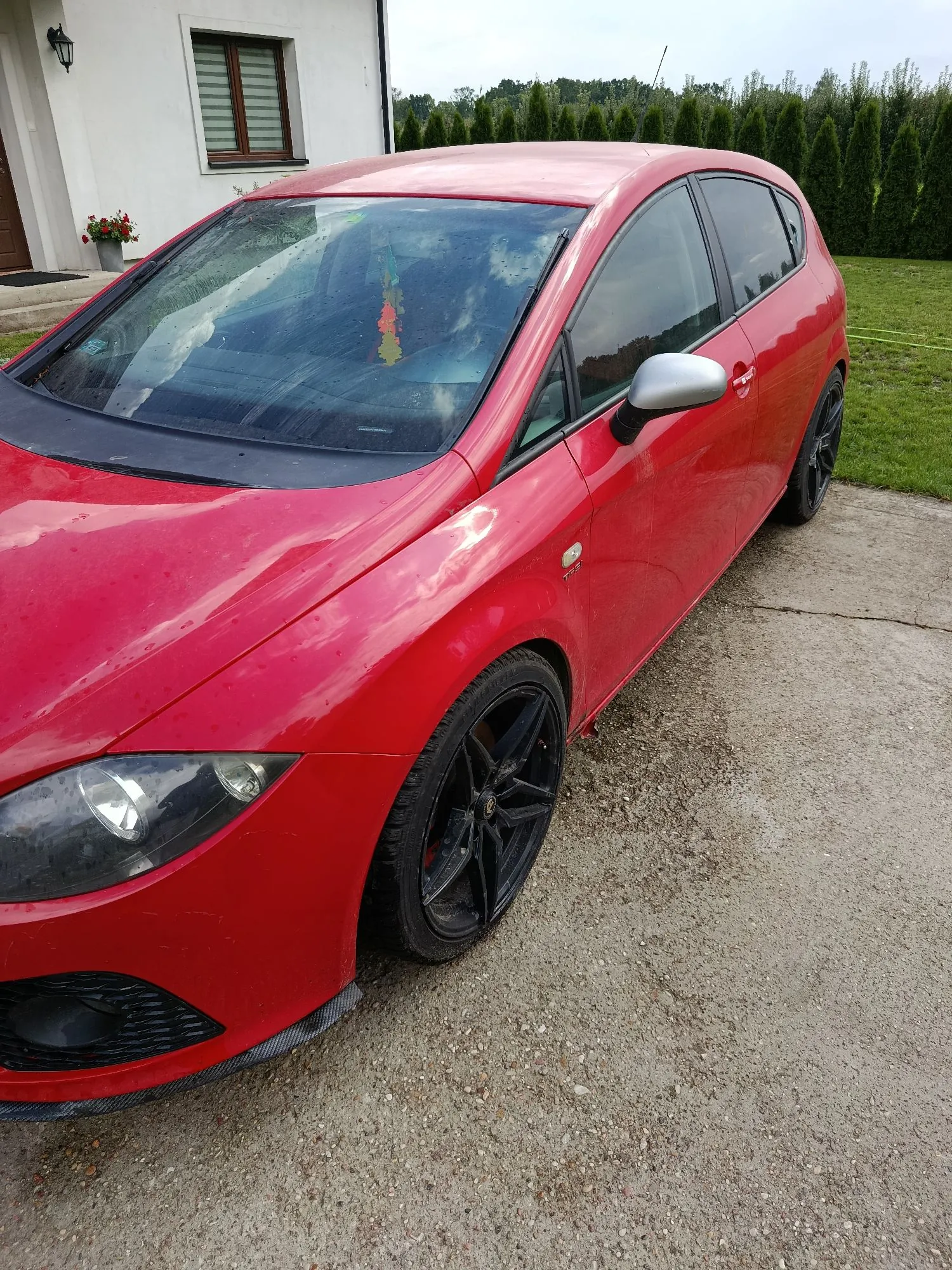 Czerwony samochód osobowy typu hatchback zaparkowany na żwirowym podjeździe przed białym budynkiem. Widoczny jest przód i bok auta, czarne felgi oraz srebrna obudowa lusterka bocznego.