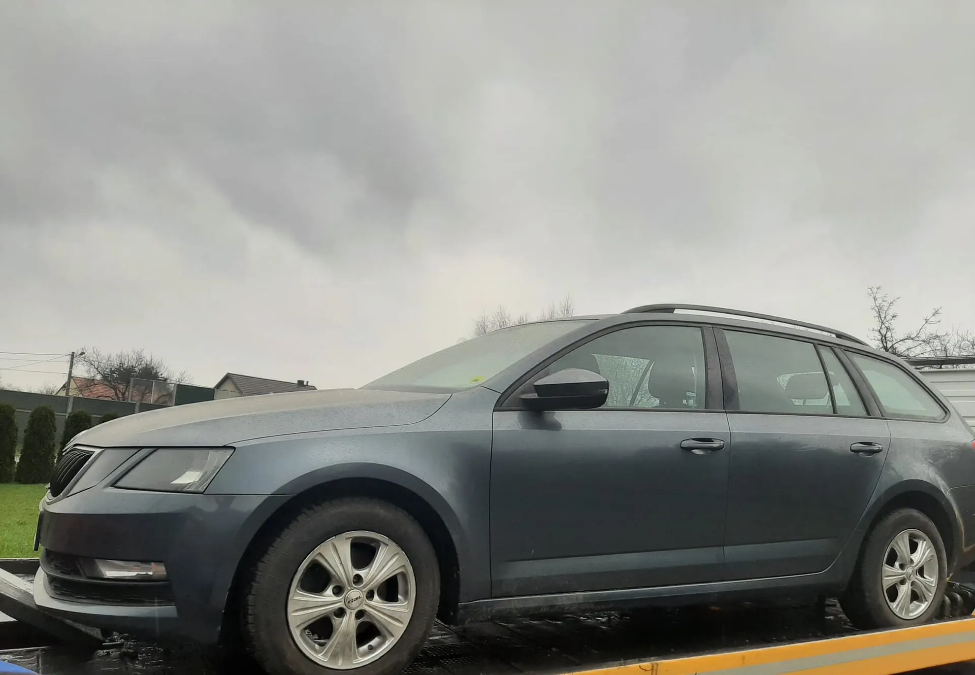 Ciemnoszary samochód typu kombi marki Škoda znajduje się na płycie holownika. Samochód ma srebrne felgi, dachowy bagażnik i jest umieszczony na czarnej powierzchni holownika z żółtą krawędzią. W tle widoczne są domy, zielona trawa, krzewy, drzewa bez liści oraz pochmurne niebo.