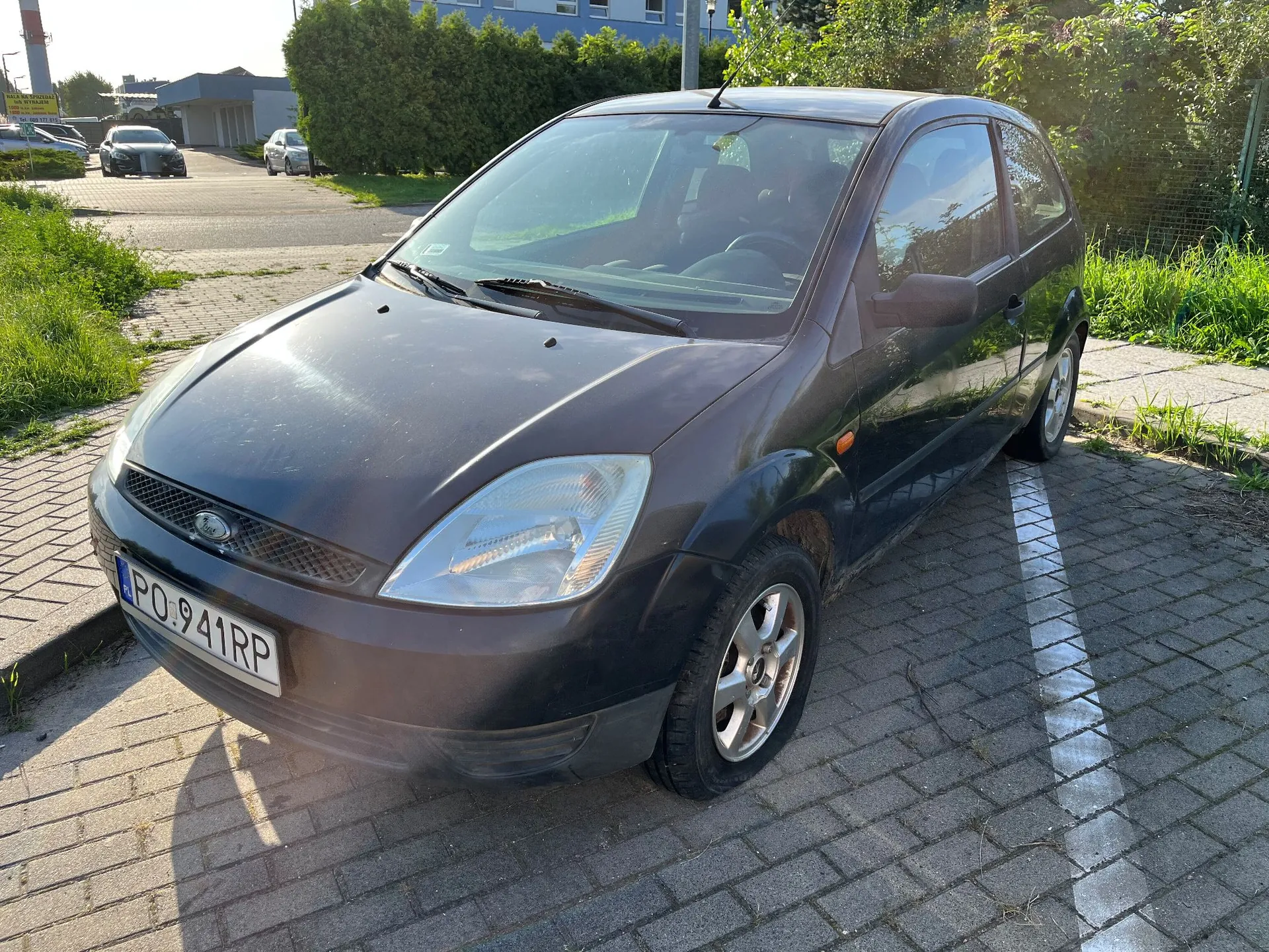 Czarny, trzy drzwiowy samochód osobowy marki Ford Fiesta zaparkowany na kostce brukowej. Pojazd ma polską tablicę rejestracyjną o numerze PO 941RP. W tle widoczne są inne samochody, zielone krzewy oraz budynki przemysłowe.