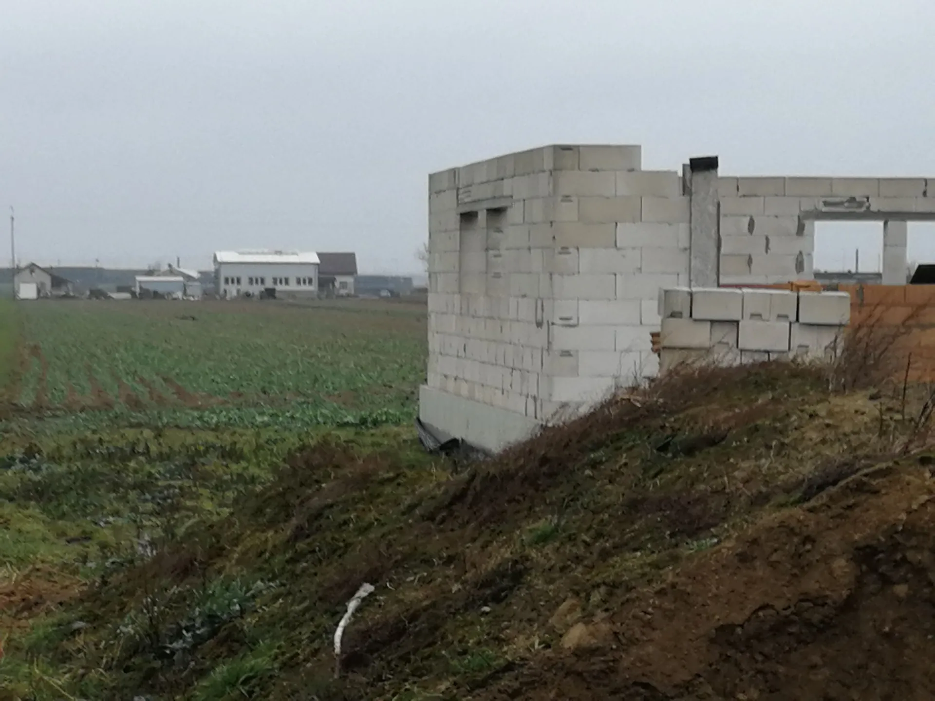 Zdjęcie przedstawia budynek w trakcie budowy, wzniesiony z jasnoszarych bloczków betonowych, znajdujący się na otwartym, trawiastym terenie. W tle widoczne są inne niskie budynki oraz szare, zachmurzone niebo. Na pierwszym planie znajduje się nasyp z ciemnej ziemi i suchej trawy.