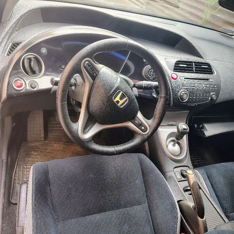Wnętrze samochodu marki Honda z perspektywy fotela kierowcy. Widoczna jest czarna kierownica z logo Hondy na środku, deska rozdzielcza z licznikiem, przyciskiem startu w kolorze czerwonym oraz konsolą centralną z nawiewami i radiem. W dolnej części zdjęcia widoczny jest fragment ciemnego, materiałowego fotela, lewarek zmiany biegów oraz hamulec ręczny.
