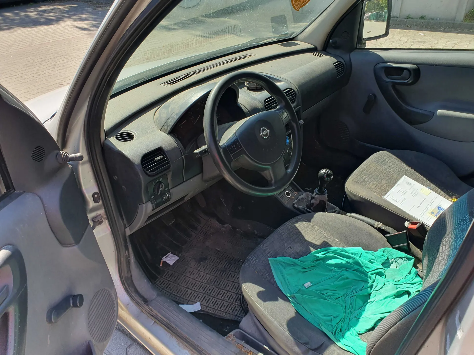 Wnętrze samochodu widziane z perspektywy otwartych przednich drzwi po stronie kierowcy. Widoczna jest deska rozdzielcza, czarna kierownica z logo Nissan, lewark zmiany biegów oraz dwa przednie fotele. Na fotelu pasażera leży biała kartka papieru, a na fotelu kierowcy znajduje się zielona tkanina. Podłoga samochodu jest zakurzona.