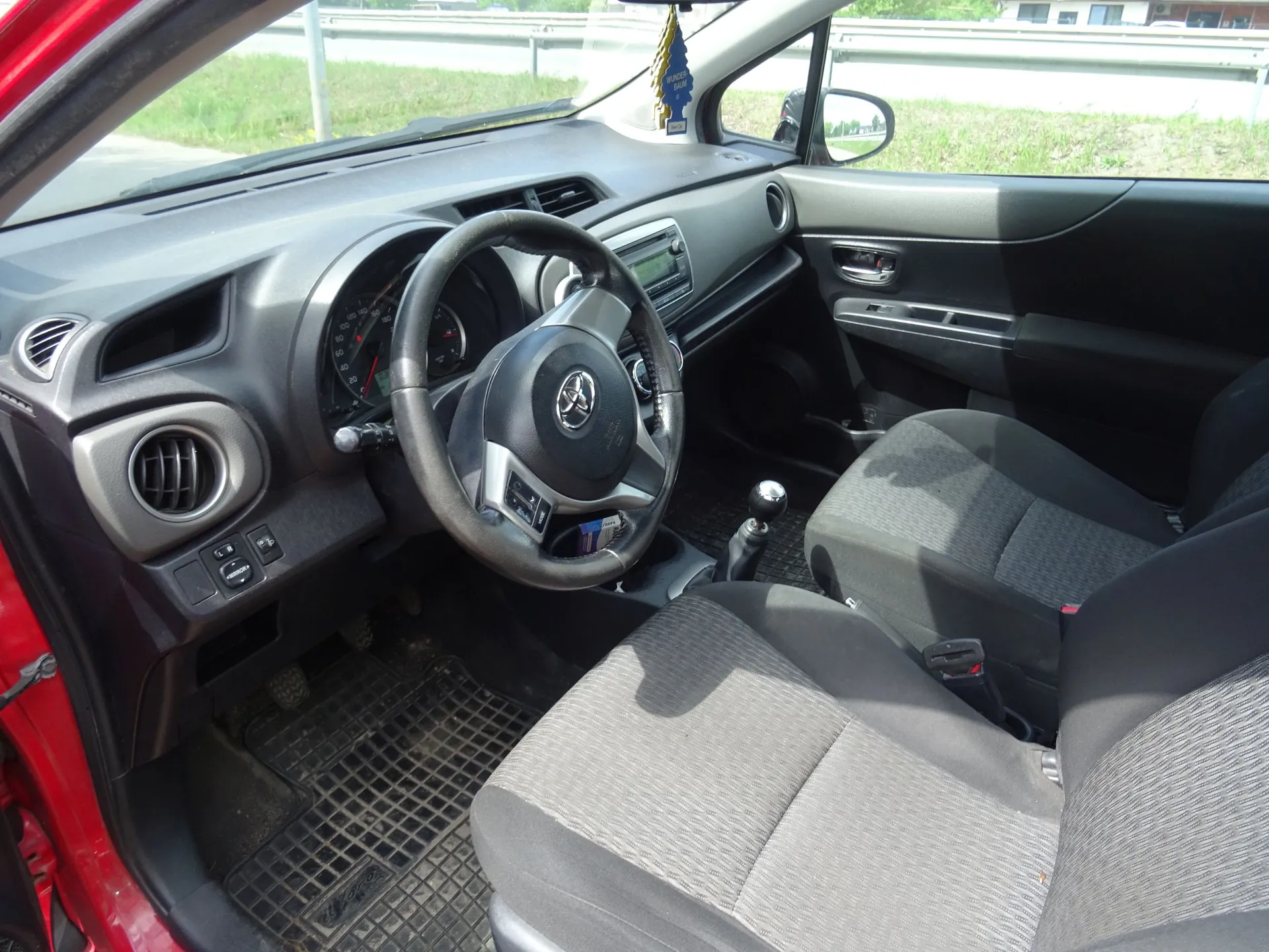 Wnętrze samochodu z perspektywy otwartych drzwi pasażera. Widoczna jest czarna deska rozdzielcza, kierownica z logo Toyoty, zegary, lewarek zmiany biegów oraz dwa przednie fotele obite ciemnoszarą tkaniną. Na podłodze znajdują się gumowe dywaniki. Samochód ma czerwony lakier zewnętrzny.