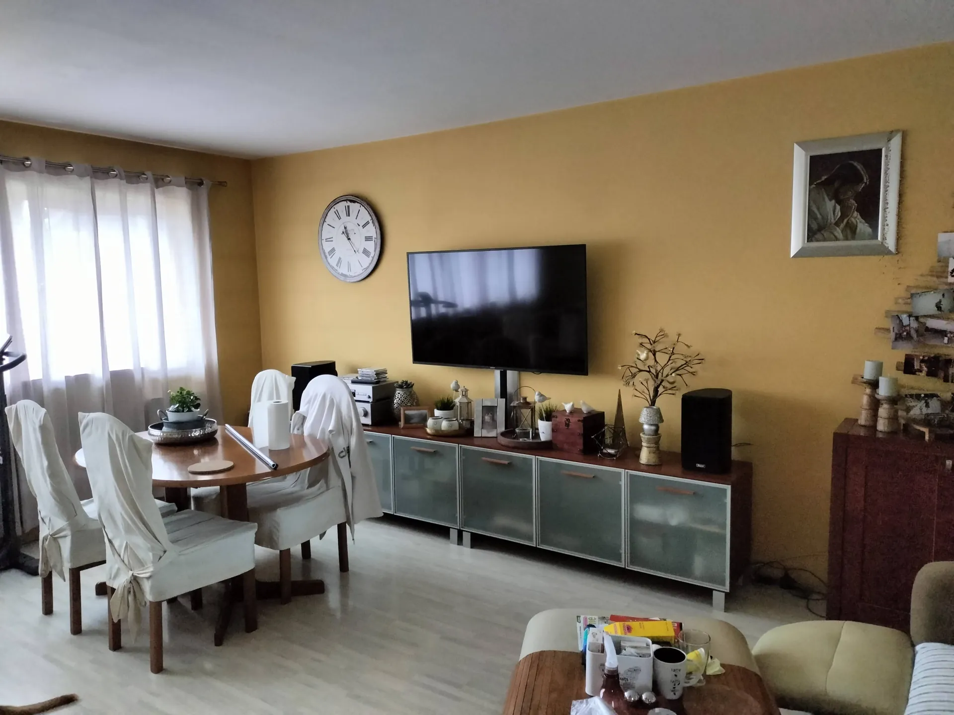 Wnętrze pokoju z żółtymi ścianami. Po lewej stronie znajduje się okno z białymi firankami, przed którym stoi drewniany stół z czterema krzesłami w białych pokrowcach. Na ścianie w centrum wisi okrągły zegar analogowy, a pod nim znajduje się długi, niski mebel RTV z szklanymi frontami, na którym stoi duży telewizor z czarnym ekranem oraz liczne dekoracje: małe rośliny, ramki ze zdjęciami i ozdobna gałązka w wazonie. Po prawej stronie widać fragment brązowej szafki oraz obraz w ramce zawieszony na ścianie. Na pierwszym planie widoczny jest fragment stolika kawowego z kubkami i przekąskami oraz fragment jasnej kanapy.