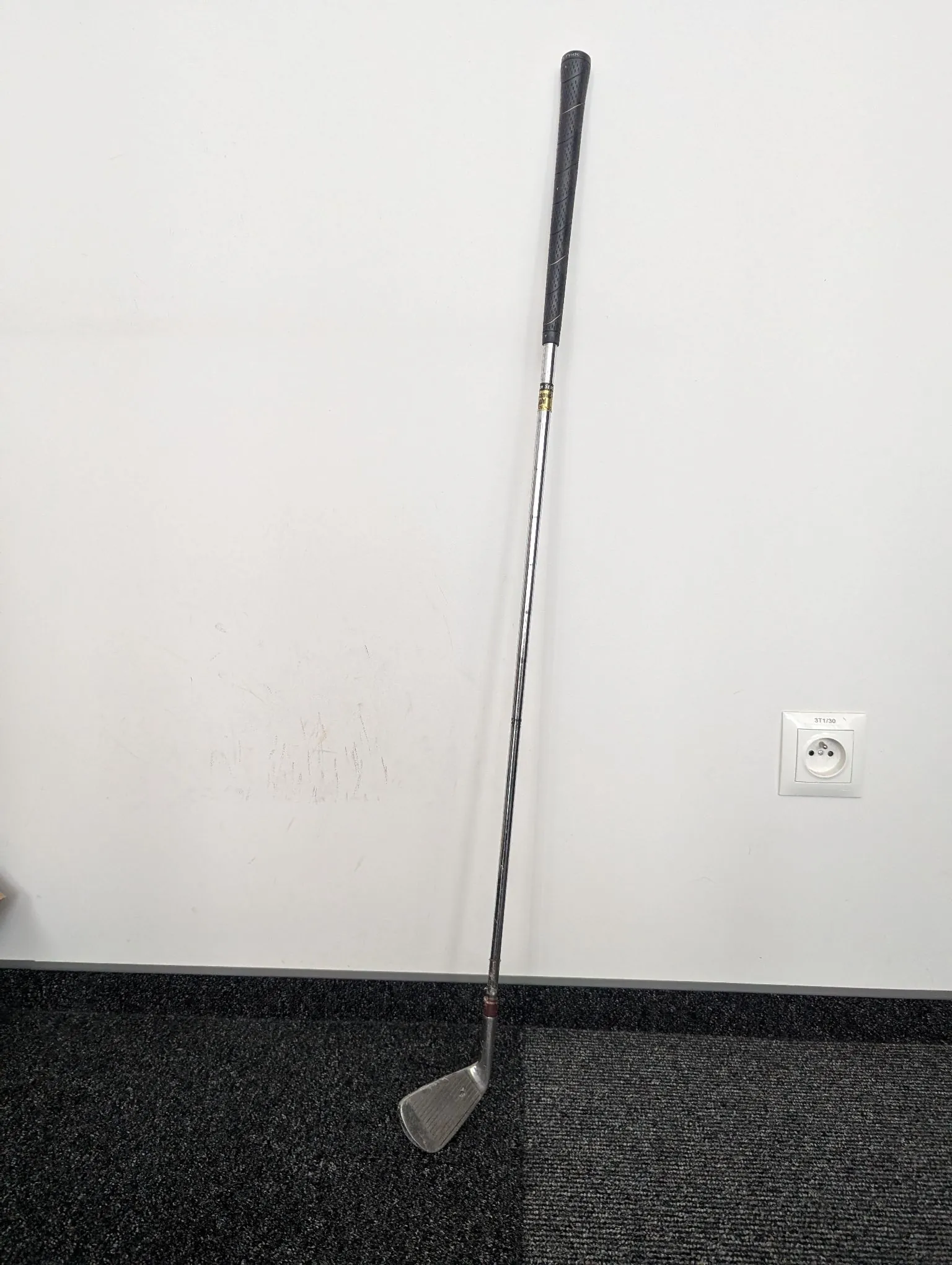 Kij golfowy oparty o białą ścianę na ciemnoszarej wykładzinie. Kij ma czarny uchwyt, metalowy trzone i srebrną główkę. Po prawej stronie na ścianie znajduje się białe gniazdko elektryczne.
