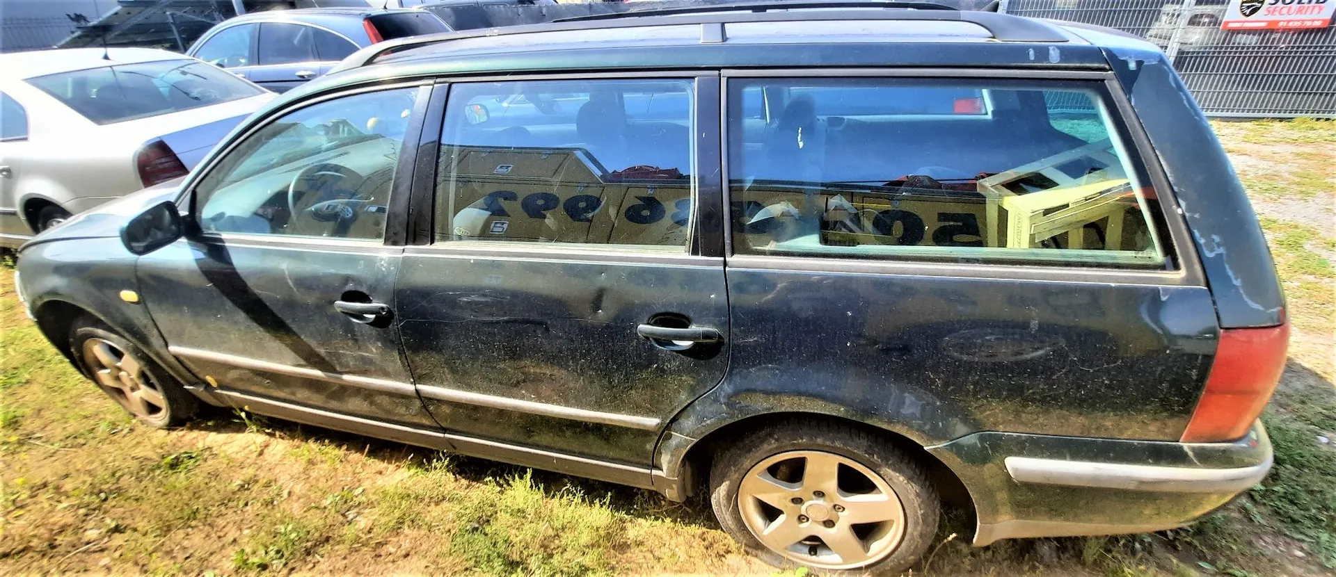 Czarny samochód typu kombi zaparkowany na trawiastym terenie. Pojazd jest brudny i posiada widoczne wgniecenia na tylnych drzwiach oraz błotniku. W tle widoczne są inne samochody oraz ogrodzenie terenu.