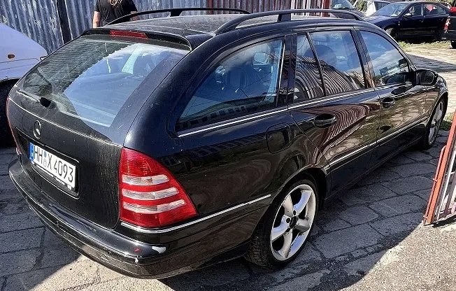 Czarny samochód marki Mercedes-Benz w nadwoziu kombi, zaparkowany na kostce brukowej. Widoczny jest tył oraz prawy bok pojazdu. Samochód posiada niemiecką tablicę rejestracyjną o numerze HH X 4093 oraz relingi dachowe. W tle widoczne są inne samochody oraz ogrodzenie.