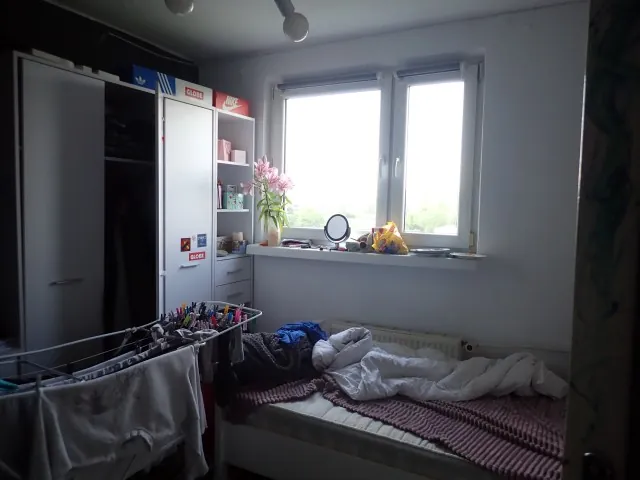 Wnętrze małego pokoju z białymi meblami. Po lewej stronie znajduje się duża biała szafa oraz węższy regał z szafkami, na których leżą pudełka marek Adidas, Globe i Nike. Na parapecie okna stoi wazon z różowymi kwiatami, małe lusterko i inne drobne przedmioty. W dolnej części obrazu widoczne jest niezasłane łóżko z białą pościelą i różowo-białym kocem. Na pierwszym planie po lewej stronie znajduje się suszarka z rozwieszonymi ubraniami.