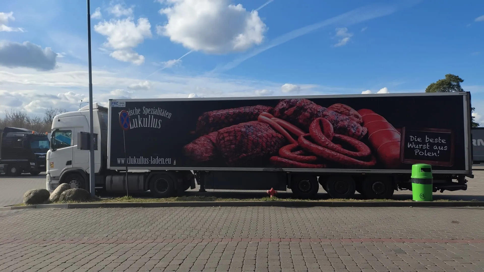 Biały ciężarówka z przyczepą z dużą reklamą przedstawiającą różne rodzaje kiełbas i mięsa. Na przyczepie widoczny napis 'Polnische Spezialitäten Lukullus', adres strony internetowej 'www.lukullus-laden.eu' oraz tabliczka z napisem 'Die beste Wurst aus Polen!'. W tle czarna ciężarówka, niebo z białymi chmurami, zielony pojemnik na śmieci oraz brukowana powierzchnia. Po lewej stronie słup i kilka kamieni.