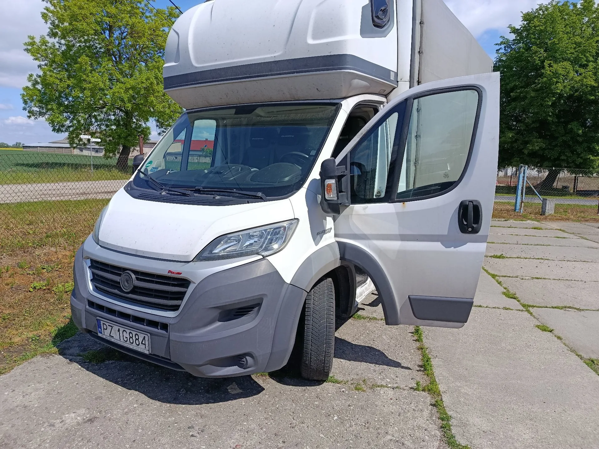 Biały samochód dostawczy typu furgon z otwartymi drzwiami kierowcy. Pojazd posiada polską tablicę rejestracyjną o numerze PZ 1G884. Auto stoi na betonowym placu, w tle widoczne są zielone drzewa i ogrodzenie.