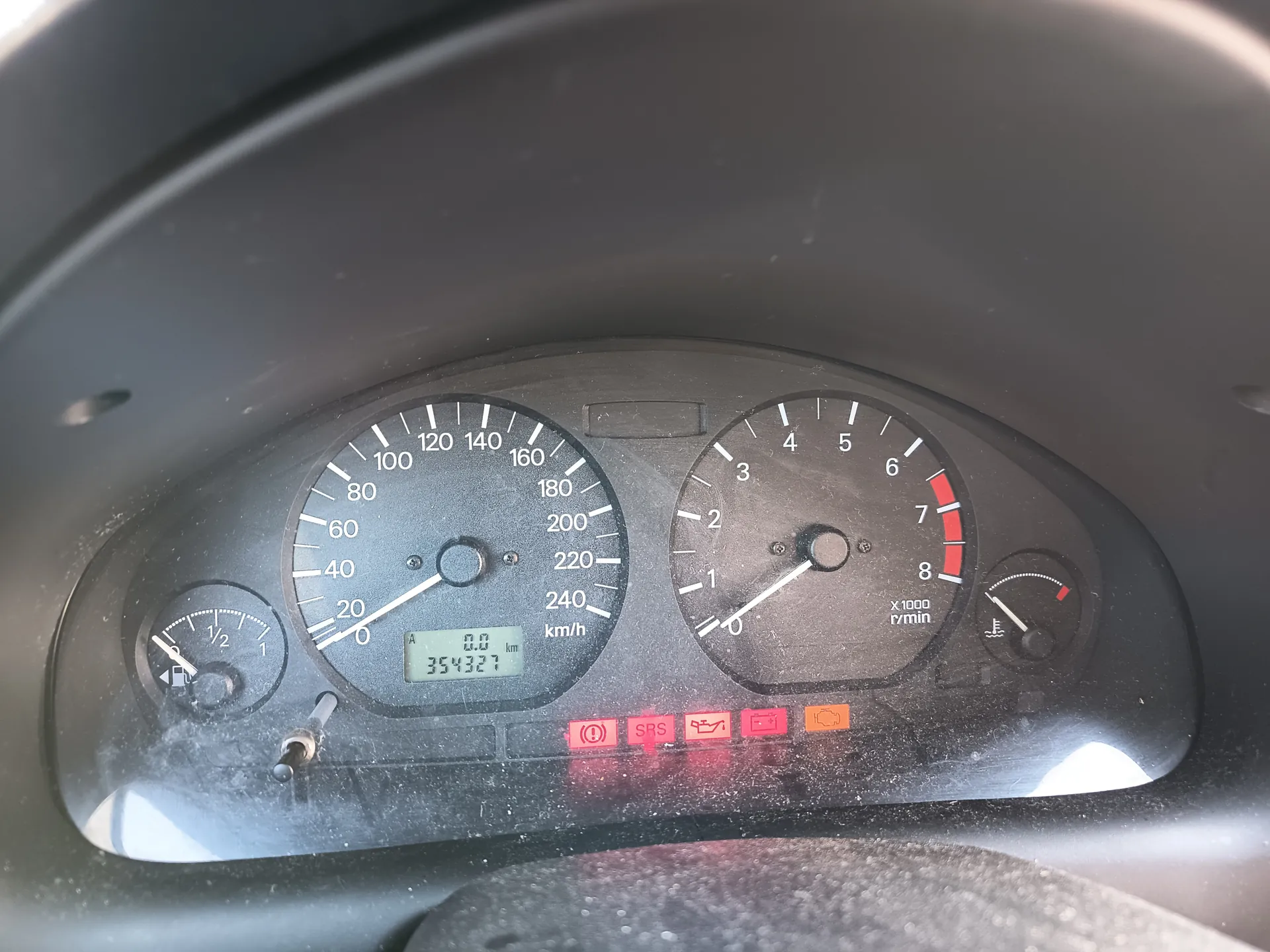 Zbliżenie na deski rozdzielcze samochodu. Po lewej stronie wskaźnik prędkości z zakresem do 240 km/h, obecna prędkość 0 km/h, licznik przebiegu 354327 km. Po prawej obrotnica z zakresem do 8 x1000 rpm, wskazanie 0. W lewym dolnym rogu wskaźnik paliwa (poziom około 1/2), w prawym dolnym wskaźnik temperatury chłodniczki. Poniżej czerwone i pomarańczowe lampki ostrzegawcze: SRS, symbol oleju, akumulatora oraz silnika. Tekst na przyrządach: km/h, x1000 r/min. Całość pokryta jest kurzem.