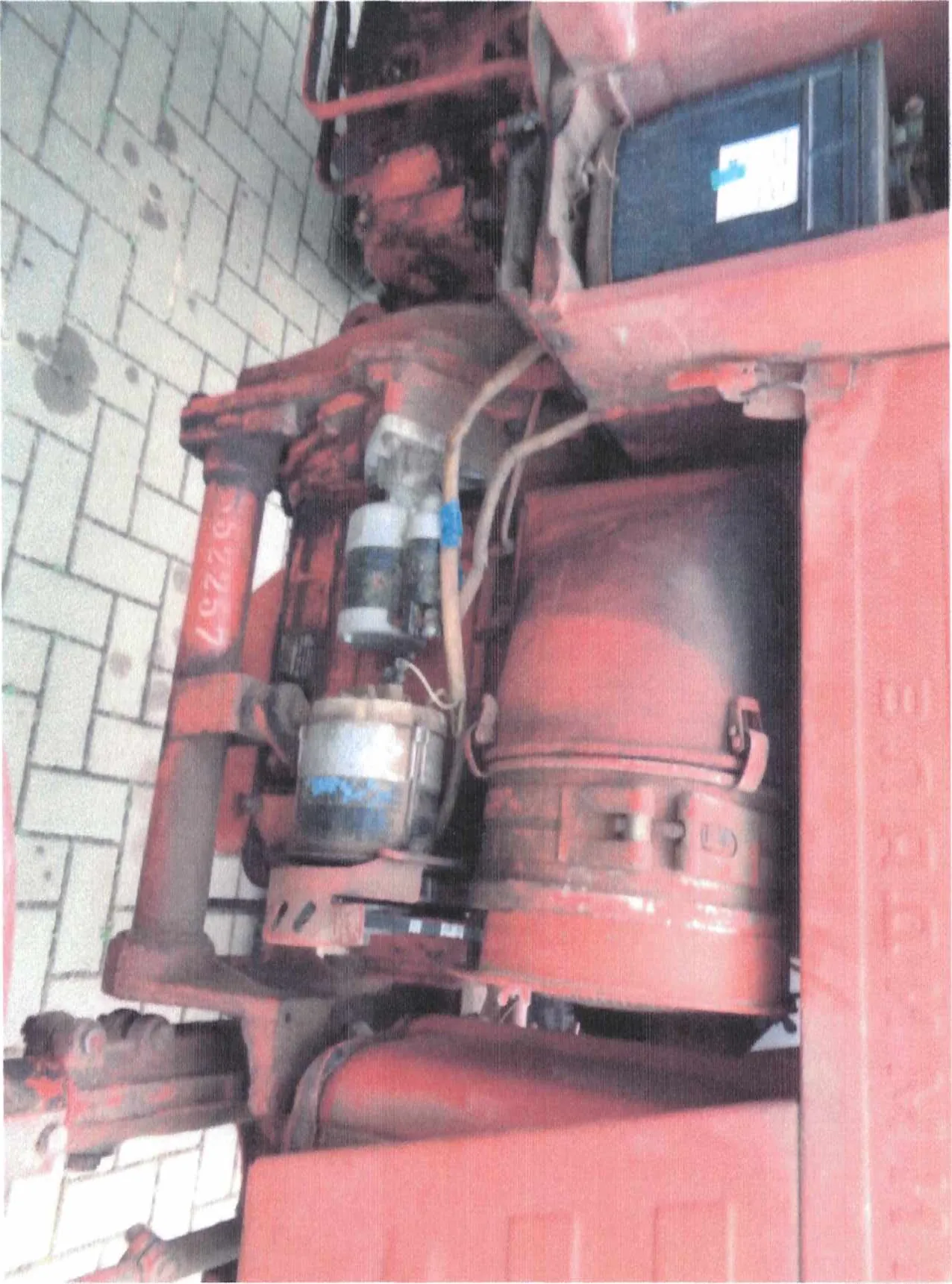 Czerwony przemysłowy element maszynowy z cylindrycznym zbiornikiem, przewodami i metalowymi częściami. Widoczny numer 552257 na czerwonej powierzchni. W tle niebieski pojemnik z białą etykietą i niebieskim znacznikiem oraz brukowana posadzka.