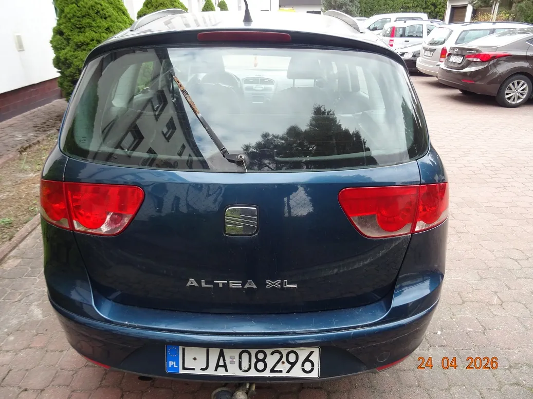 Tylny widok ciemnoniebieskiego samochodu marki Seat Altea XL. Na klapie bagażnika znajduje się logo marki oraz napis 'ALTEA XL'. Samochód ma polską tablicę rejestracyjną o numerze LJA 08296. W prawym dolnym rogu zdjęcia widnieje data 24 04 2026. W tle widoczne są inne zaparkowane samochody oraz zielone krzewy.