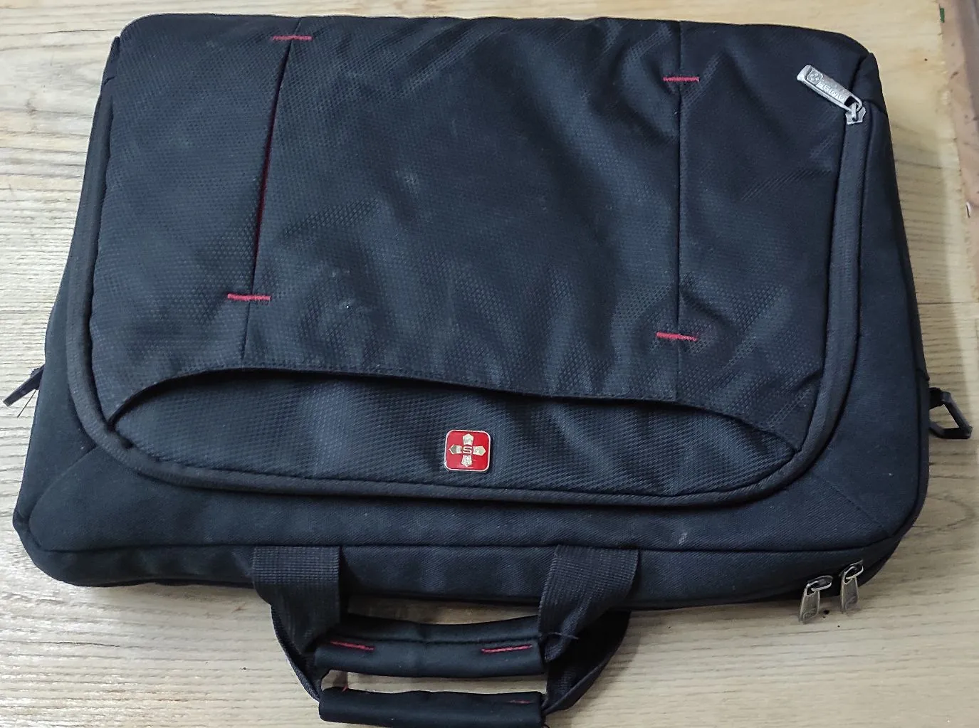 Czarna torba na laptopa leżąca na drewnianej powierzchni. Torba ma główną klapę z zaokrąglonym dołem, na której znajduje się małe, czerwono-białe logo w kształcie tarczy. Widoczne są czerwone przeszycia w kilku miejscach oraz dwa uchwyty do przenoszenia na dole obrazu.