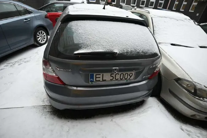 Zdjęcie przedstawia szary samochód Honda z tyłu, zaparkowany na śnieżnej powierzchni. Na tylnej szybie i dachu widoczny jest śnieg. Tablica rejestracyjna oznaczona jako EL 5C009 z flagą Polski po lewej stronie. Po obu stronach samochodu znajdują się inne pojazdy: niebieski po lewej i beżowy po prawej, również pokryte śniegiem. W tle widoczne są budynki z oknami oraz osoba stojąca za samochodem. Całość otoczona jest śniegiem na ziemi.