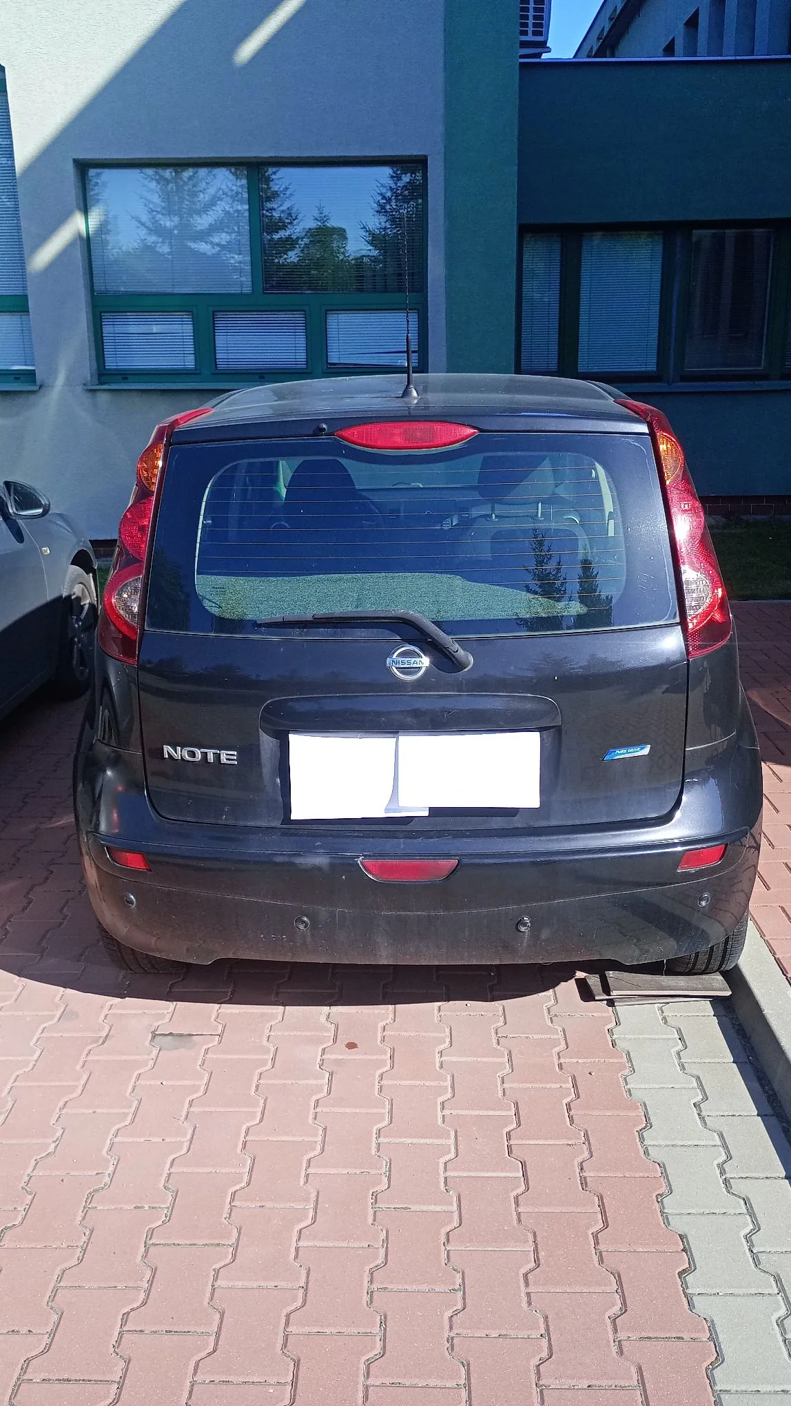 Tył ciemnoszarego samochodu marki Nissan Note zaparkowanego na kostce brukowej przed budynkiem. Na klapie bagażnika widoczne jest logo Nissana, napis NOTE oraz zasłonięta biała tablica rejestracyjna. Samochód ma tylną szybę z wycieraczką i światła stop po bokach.