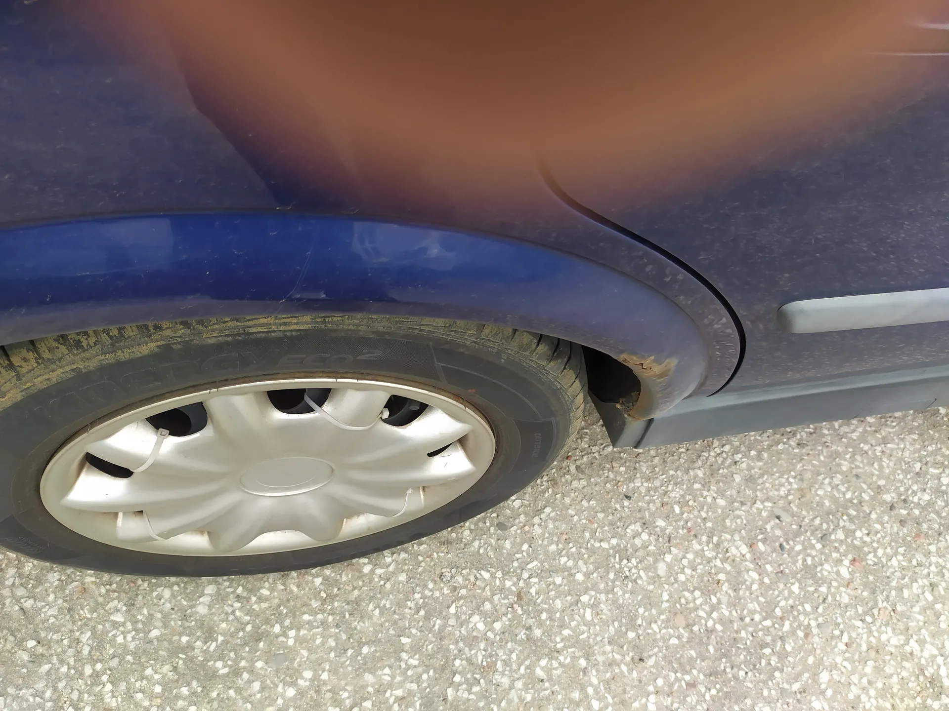 Zbliżenie na przednie koło niebieskiego samochodu. Widoczna opona z napisem ENERGY ECO 2, srebrna felga oraz fragment błotnika z widocznymi śladami rdzy. Samochód stoi na nawierzchni z drobnych kamieni i asfaltu.