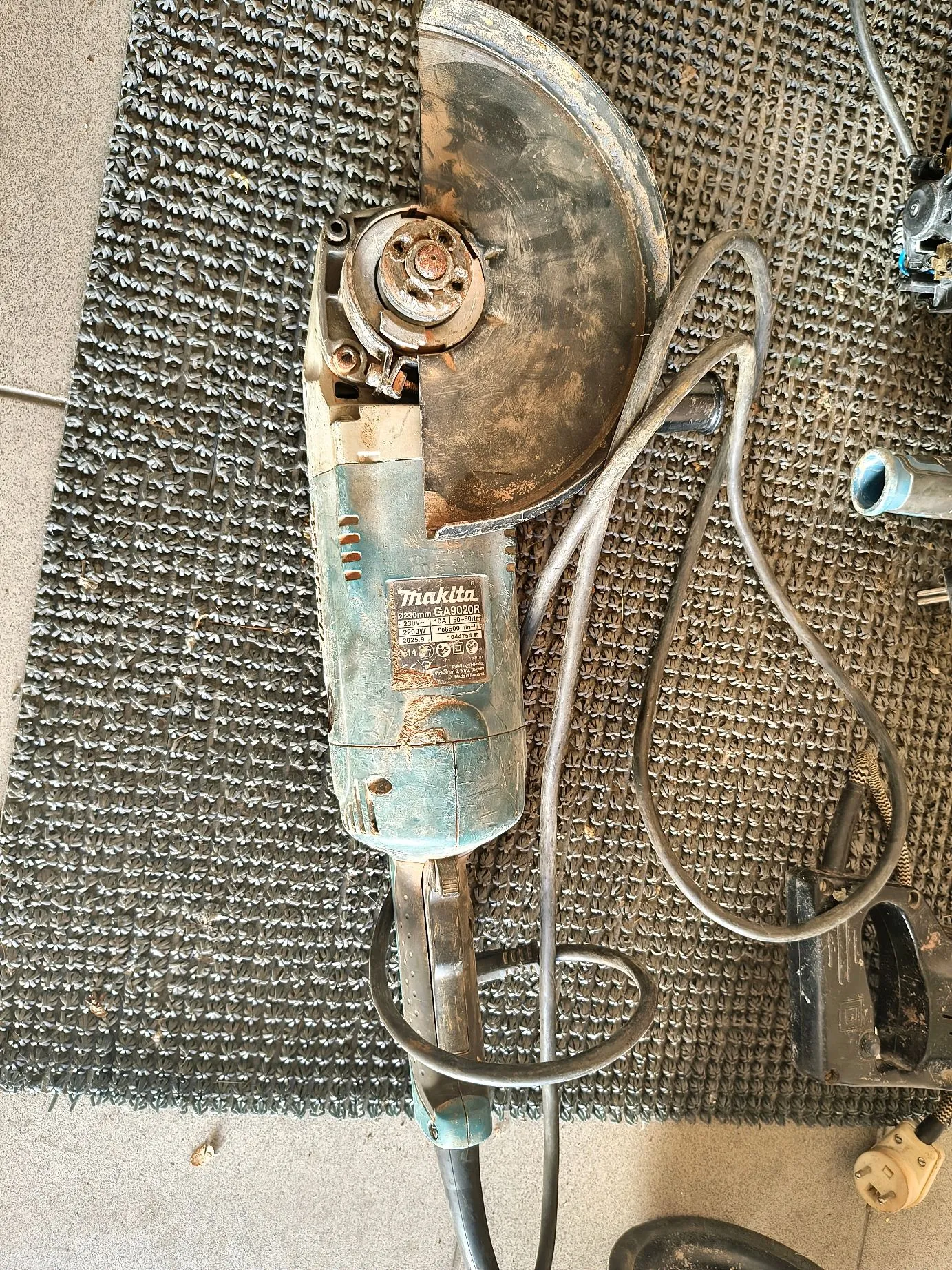 Szarozielona szlifierka kątowa marki Makita, model GA9020R, leżąca na szarej, teksturowanej macie. Urządzenie jest zabrudzone pyłem budowlanym. Widoczna jest metalowa osłona tarczy oraz czarny kabel zasilający owinięty wokół narzędzia.