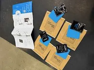 Na ciemnym tle widoczne są cztery brązowe pudełka z niebieskimi etykietami. Na każdym pudełku leży czarny elektroniczny komponent. Po lewej stronie znajduje się biały arkusz z instrukcją zawierający schematy i nieczytelny tekst. W tle widoczna jest część jasnej podłogi.