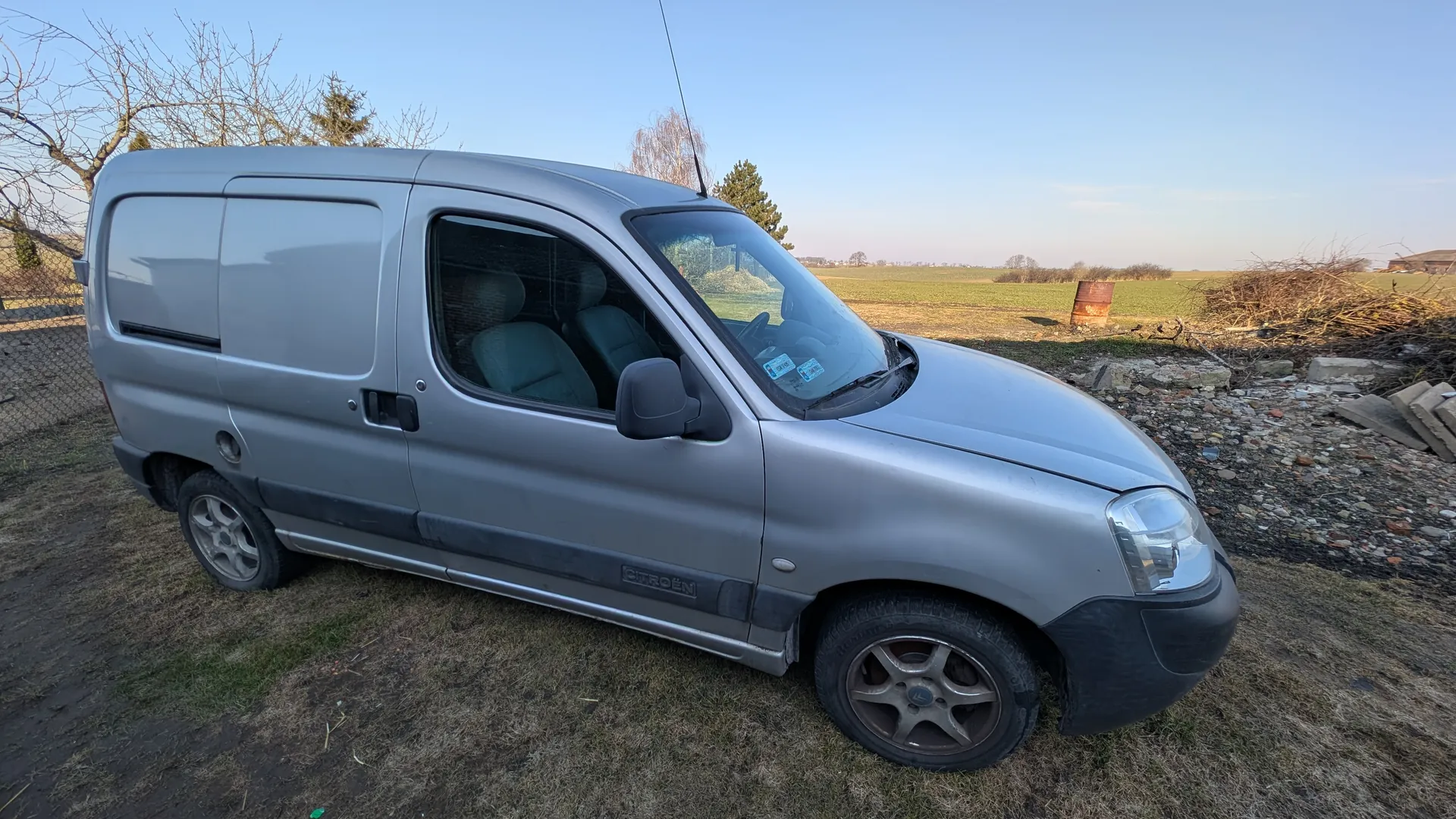 Srebrny samochód dostawczy Citroën zaparkowany na trawiastym terenie z widocznymi śladami błota. Po bokach pojazdu znajdują się drzwi z oknem oraz zasłonięte tylne okna. W tle rozległe pole, suche drzewa, rdzewiejąca beczka i stos gałęzi. Niebo jest jasne i bezchmurne. Na boku pojazdu widoczny napis "CITROËN".