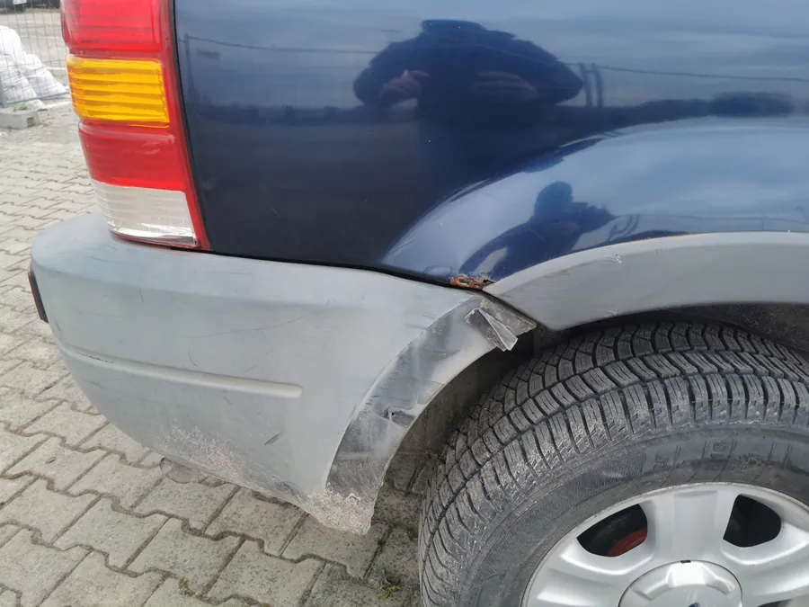 Zdjęcie przedstawia tylną część ciemnoniebieskiego samochodu, konkretnie prawą stronę z widocznym tylnym światłem (czerwone i żółte sekcje), uszkodzonym szarym zderzakiem z widocznymi zadrapaniami i rdzą oraz kołem z srebrną felgą. Na karoserii odbija się sylwetka osoby robiącej zdjęcie. Samochód stoi na brukowanej powierzchni z kostki betonowej. W tle widoczny jest ogrodzenie i otwarta przestrzeń.