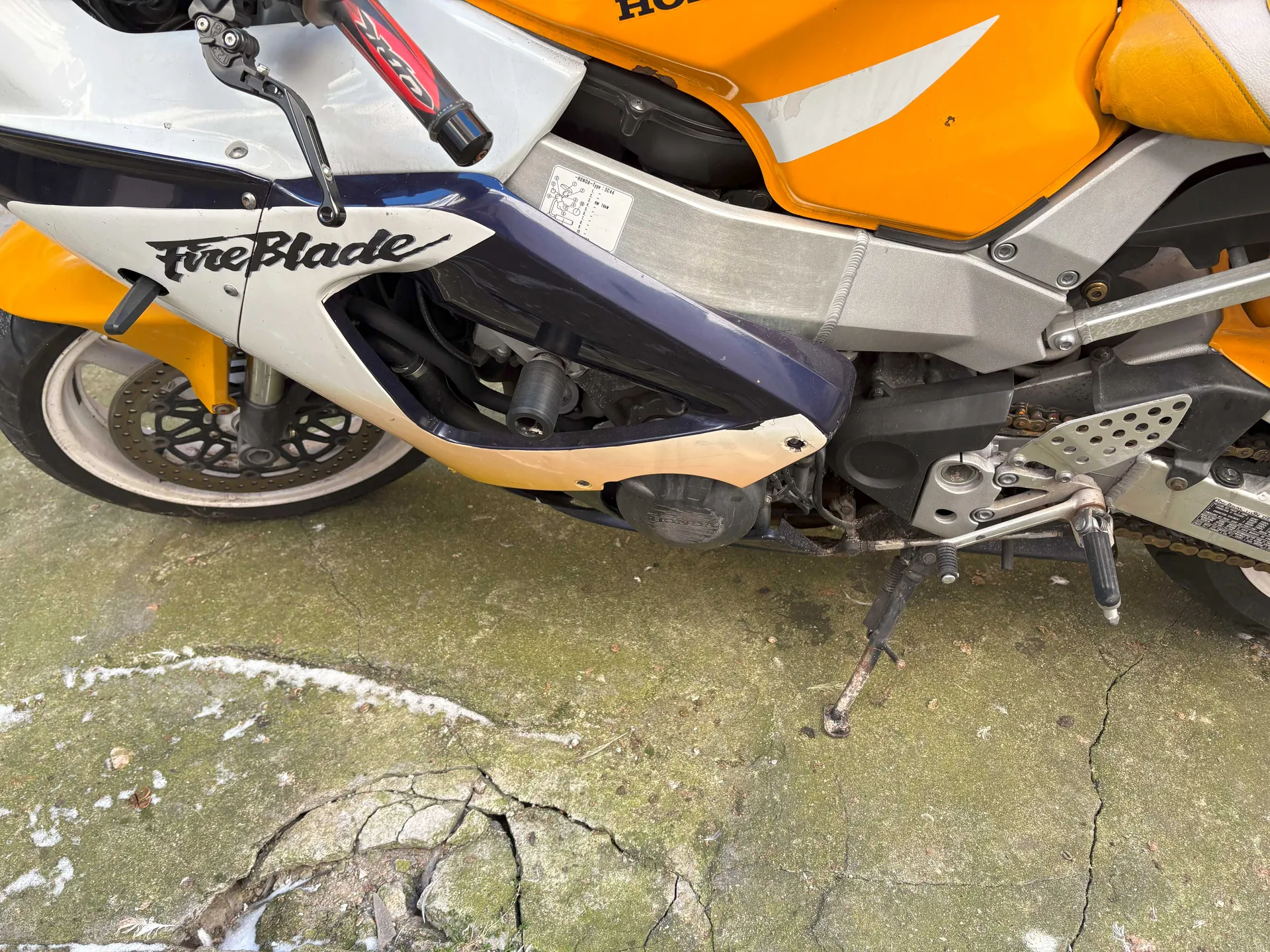 Zbliżenie na fragment motocykla marki Honda z widocznym napisem 'FireBlade' na białym i ciemnoniebieskim nadkolu. Motocykl ma pomarańczowe i srebrne elementy, widoczne są przednie koło z tarczą hamulcową, łańcuch, podstawa i fragmenty mechanizmów. Pojazd stoi na pękniętej betonowej powierzchni z resztkami śniegu lub bieli. Na nadkolu znajduje się również częściowo widoczny napis 'HONDA'.