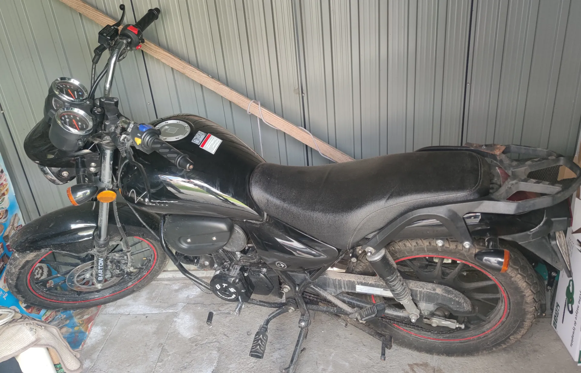 Czarny motocykl z czerwonymi akcentami na felgach, zaparkowany przed szarą blachą falowaną. Motocykl posiada czarne siedzenie, widoczne wskaźniki prędkości i obrotomierze, kierownicę z przyciskami oraz tabliczkę z napisem "BARTON" na przednim widelcu. Na silniku widoczny napis "DTS". Po lewej stronie obrazu częściowo widoczny kolorowy plakat lub gazeta na betonowej podłodze, po prawej biała pudełko z zielonym logo. W tle oparta o ścianę drewniana belka. Motocykl oparty na podstawce.