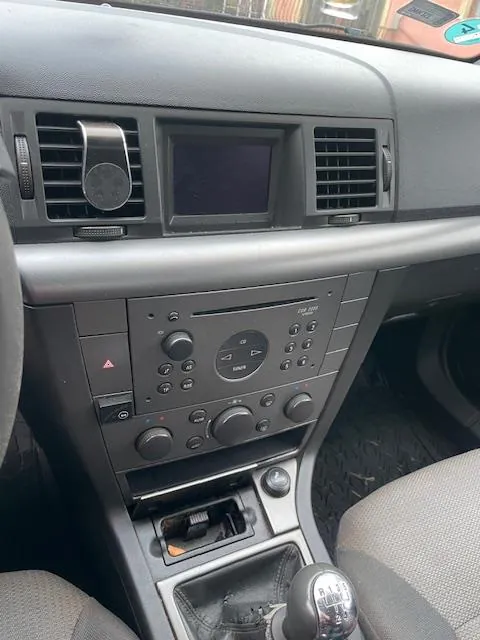 Wnętrze samochodu z widokiem na deski rozdzielczą. Widoczne są dwa wentylatory powietrza po bokach, centralny ekran, panel sterowania z przyciskami i pokrętłami, w tym oznaczeniami takimi jak 'P R N D 3 2 1' na dźwigni biegów. Siedzenia są tapicerowane, a podłoga pokryta jest czarną wykładziną. W tle przez przednią szybę widać fragmenty otoczenia.