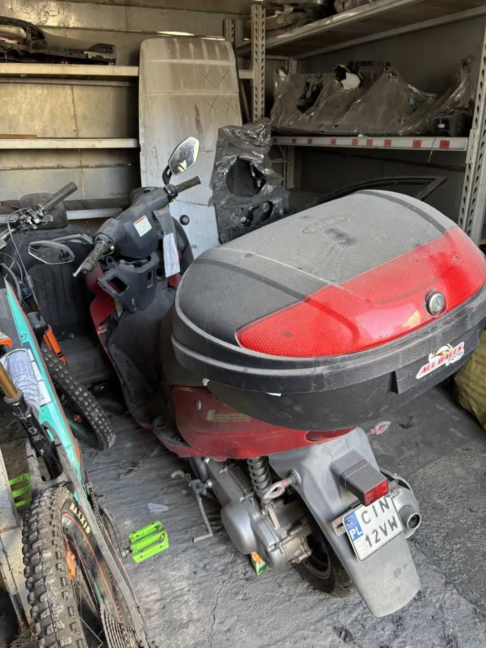 Czerwono-czarny skuter z dużym bagażnikiem, tablicą rejestracyjną PL CIN 12VW, stojący w garażu obok turkusowego roweru z grubymi oponami. W tle metalowe półki z samochodowymi częściami, w tym siedzeniami i elementami zakrytymi folią. Podłoga z betonu, na niej widoczne zielone elementy i śmieci.