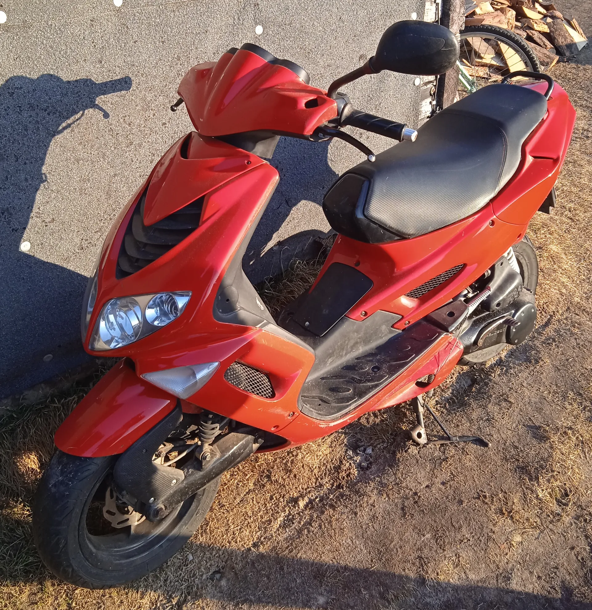 Czerwony skuter stojący na suchym trawniku i asfalcie. Skuter ma czarne siedzenie, reflektory, lusterka boczne i widoczne elementy mechaniczne. W tle widoczne są fragmenty koła roweru oraz stos drewna. Na asfalcie widoczny jest cień osoby trzymającej urządzenie do robienia zdjęć.