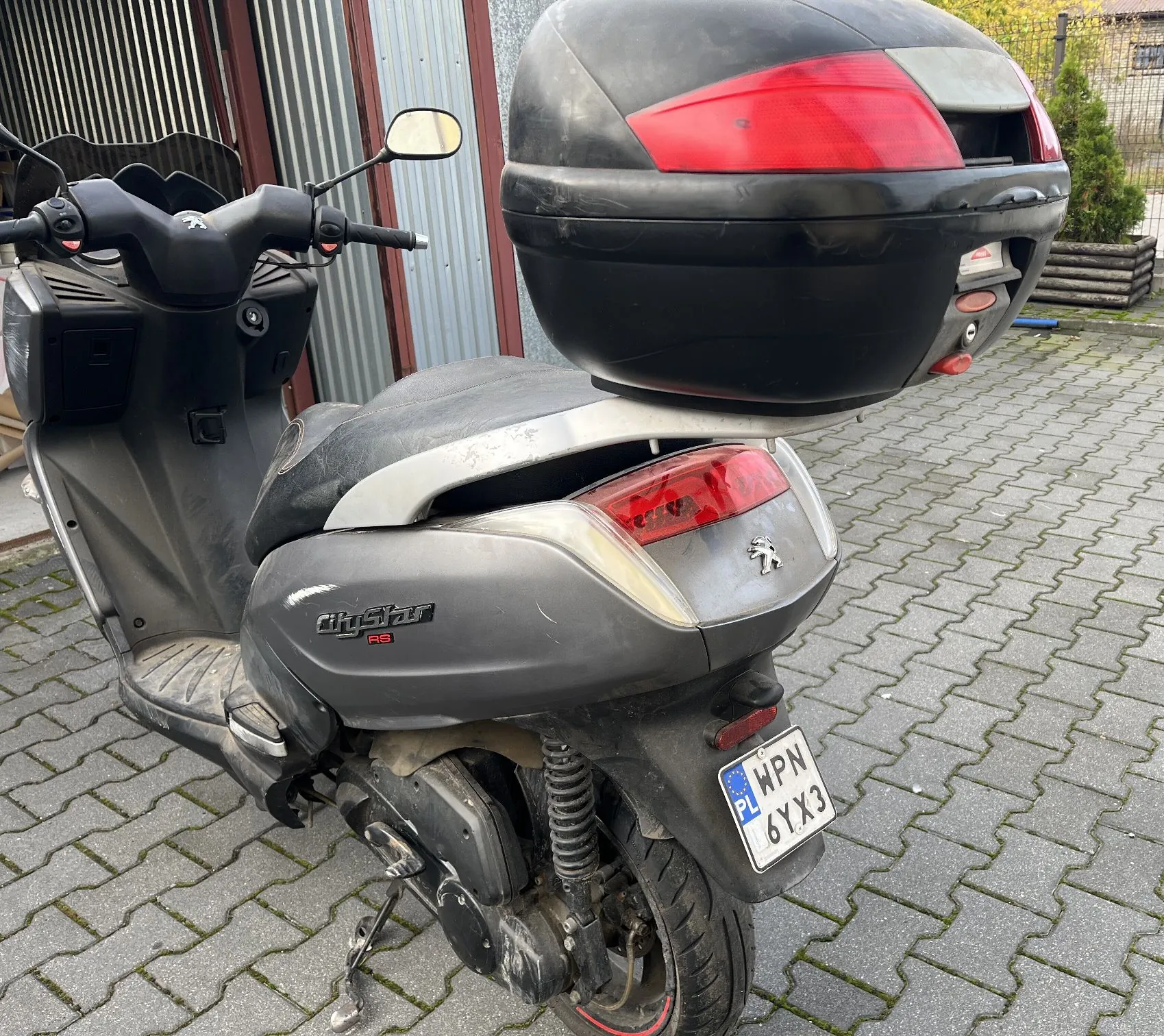 Szary skuter marki Peugeot model CityStar RS zaparkowany na kostce brukowej. Pojazd posiada zamontowany czarny kufer tylny. Widoczna jest tablica rejestracyjna o numerze WPN 6YX3 z oznaczeniem PL.