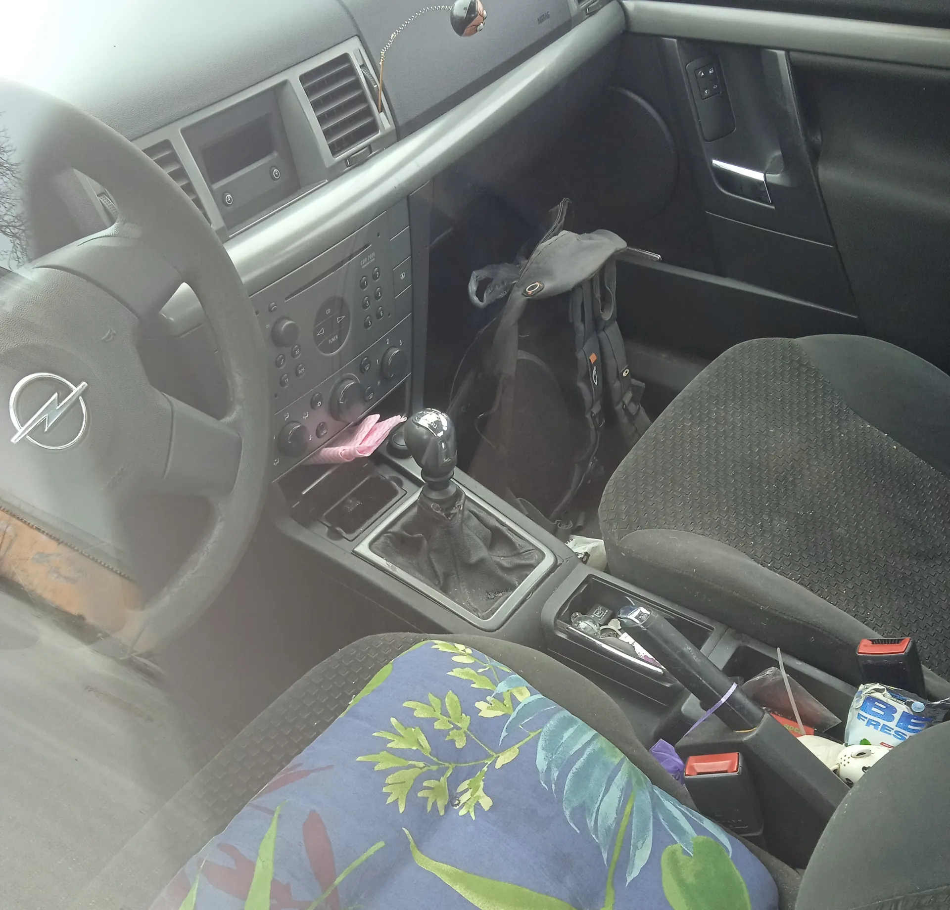 Wnętrze samochodu z widocznym kierownicą z logo Opel po lewej stronie, deska rozdzielcza z radioodtwarzaczem, przyciskami i wentylacją, dźwignią zmiany biegów w środku, czarnym plecakiem na pasażerskim siedzeniu, kolorową poduszką na siedzeniu kierowcy z niebieskim tłem i zielonymi liśćmi, różową tkaniną na konsoli środkowej oraz opakowaniem z napisem BE FRESH w przestrzeni na przednim siedzeniu. Widoczne są również uchwyty drzwiowe i sterowanie oknami.