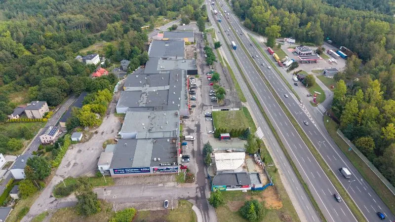 Widok z lotu ptaka na obszar przemysłowo-handlowy położony obok wielopasmowej drogi ekspresowej. Po lewej stronie znajduje się kompleks dużych, szarych hal magazynowych i produkcyjnych z płaskimi dachami. Przed halami widoczne są parkingi z zaparkowanymi samochodami osobowymi. W oddali po lewej stronie widać domy jednorodzinne otoczone gęstym lasem. Po prawej stronie przebiega szeroka droga z kilkoma pasami ruchu w obu kierunkach, na której poruszają się samochody i ciężarówki. Przy drodze znajduje się stacja paliw z białym zadaszeniem i obszarem serwisowym. Całość otoczona jest zielenią i drzewami.