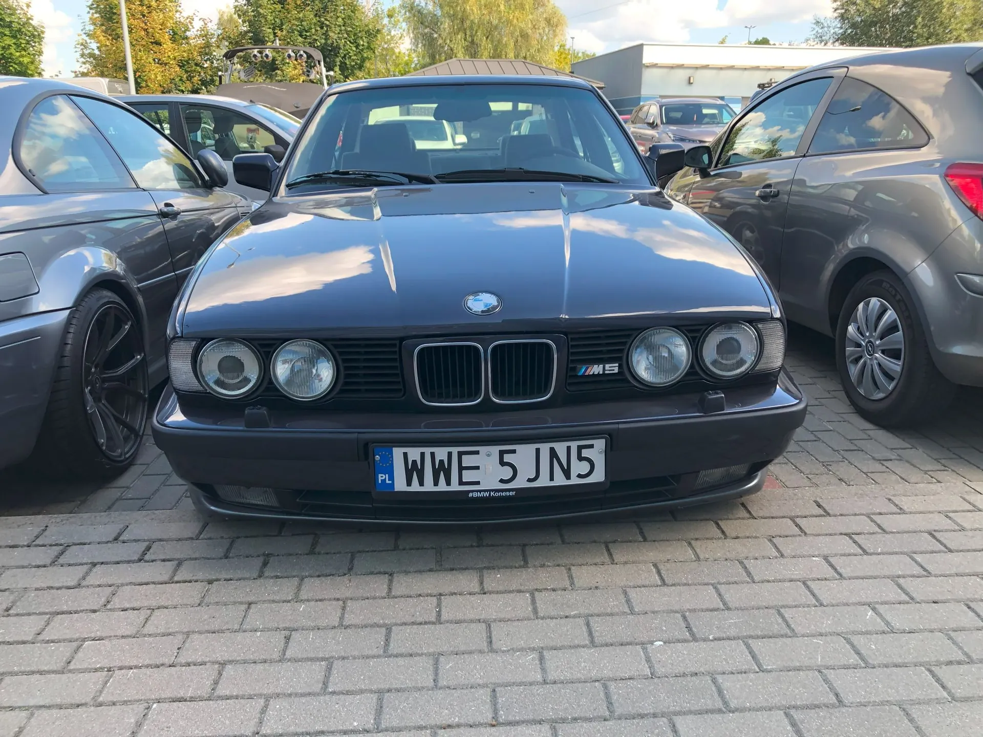 Zdjęcie przedstawia przód ciemnoszarego samochodu BMW serii 5 (E34) z logo M5 na grillu. Samochód posiada polską tablicę rejestracyjną o numerze WWE 5JN5. Pojazd zaparkowany jest na kostce brukowej pomiędzy dwoma innymi samochodami w kolorze szarym. W tle widoczne są drzewa i budynki przemysłowe pod jasnym niebem.