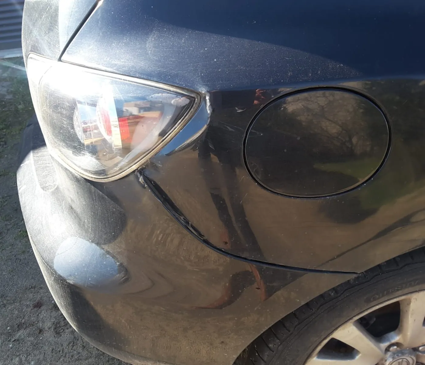 Zbliżenie przedniej części ciemnoszarego samochodu. Widoczny jest reflektor z odbiciem budynków i drzew, uszkodzony zarysowany zarys na błotniku oraz koło z oponą z napisem OUTSIDE na boku. Na karoserii widoczne są ślady zabrudzeń i odbicia otoczenia.