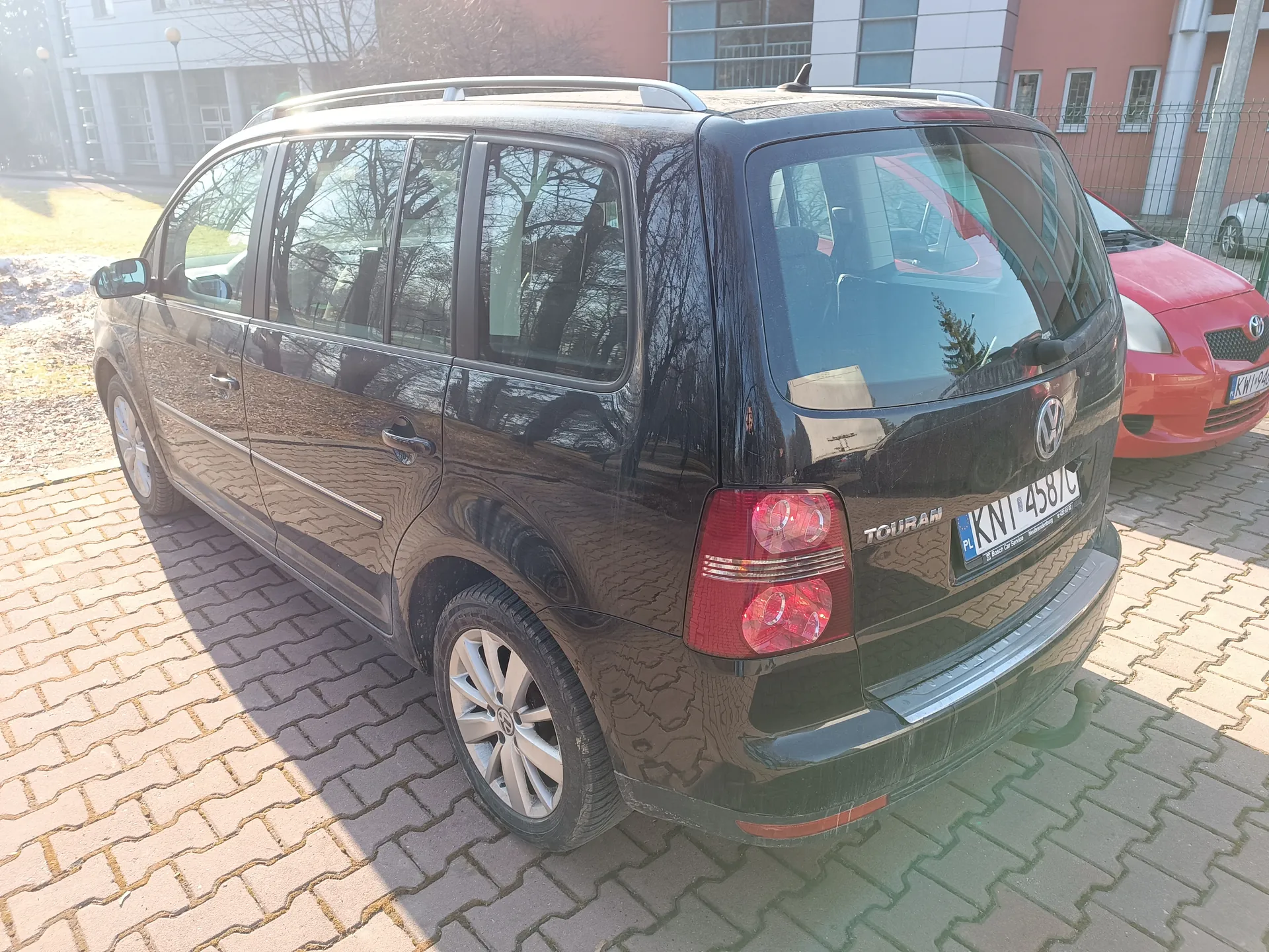Czarny samochód Volkswagen Touran zaparkowany na brukowanym placu. Widoczny tył pojazdu z tablicą rejestracyjną PL KNT 458 C oraz napisem TOURAN. Obok, po prawej stronie, czerwony samochód Toyota. Tło stanowią budynki z oknami, ogrodzenie i drzewa bez liści. Na ziemi fragmenty śniegu i sucha trawa.