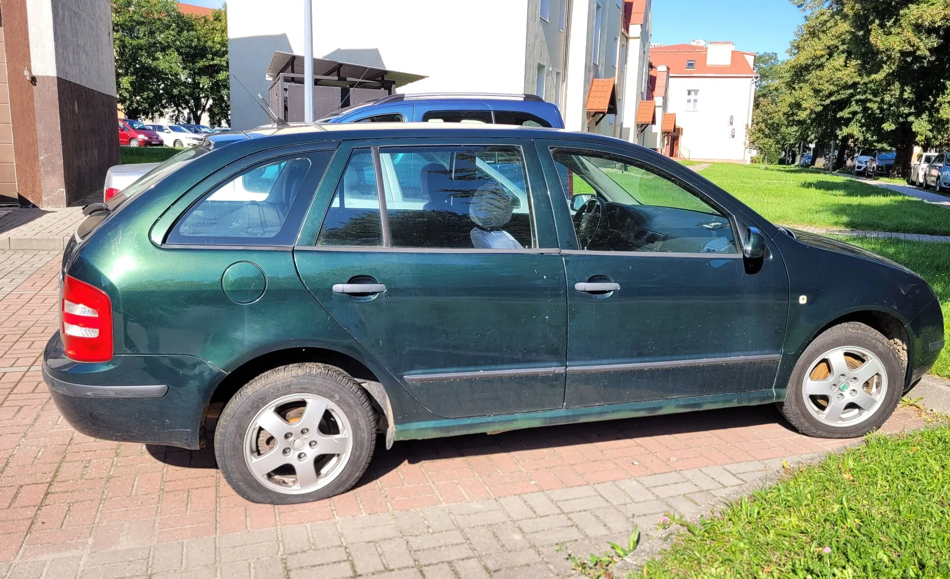 Ciemnozielony samochód osobowy typu kombi zaparkowany na kostce brukowej. Pojazd jest pokazany z profilu od strony pasażera. W tle widoczne są białe budynki mieszkalne, zielone drzewa oraz fragment innego, niebieskiego samochodu.