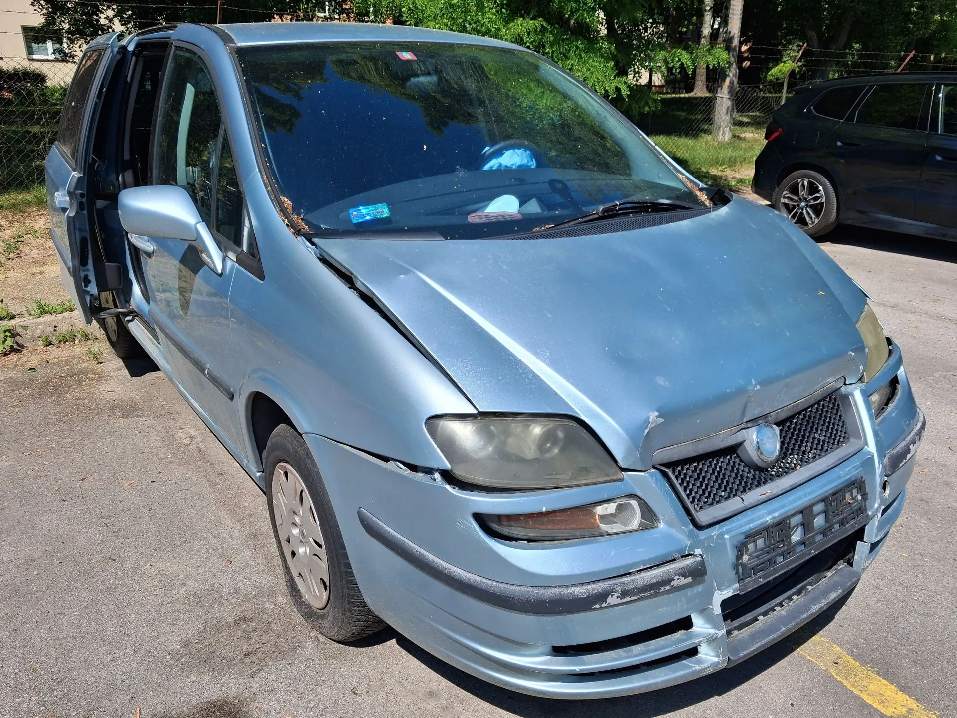 Jasnoniebieski samochód typu minivan z widocznymi uszkodzeniami karoserii. Przednia część pojazdu jest wgnieciona, maska jest podniesiona i zdeformowana, a przedni zderzak jest pęknięty i częściowo oderwany. Lewe przednie drzwi są otwarte. Samochód stoi na asfaltowym parkingu na tle zielonych drzew.