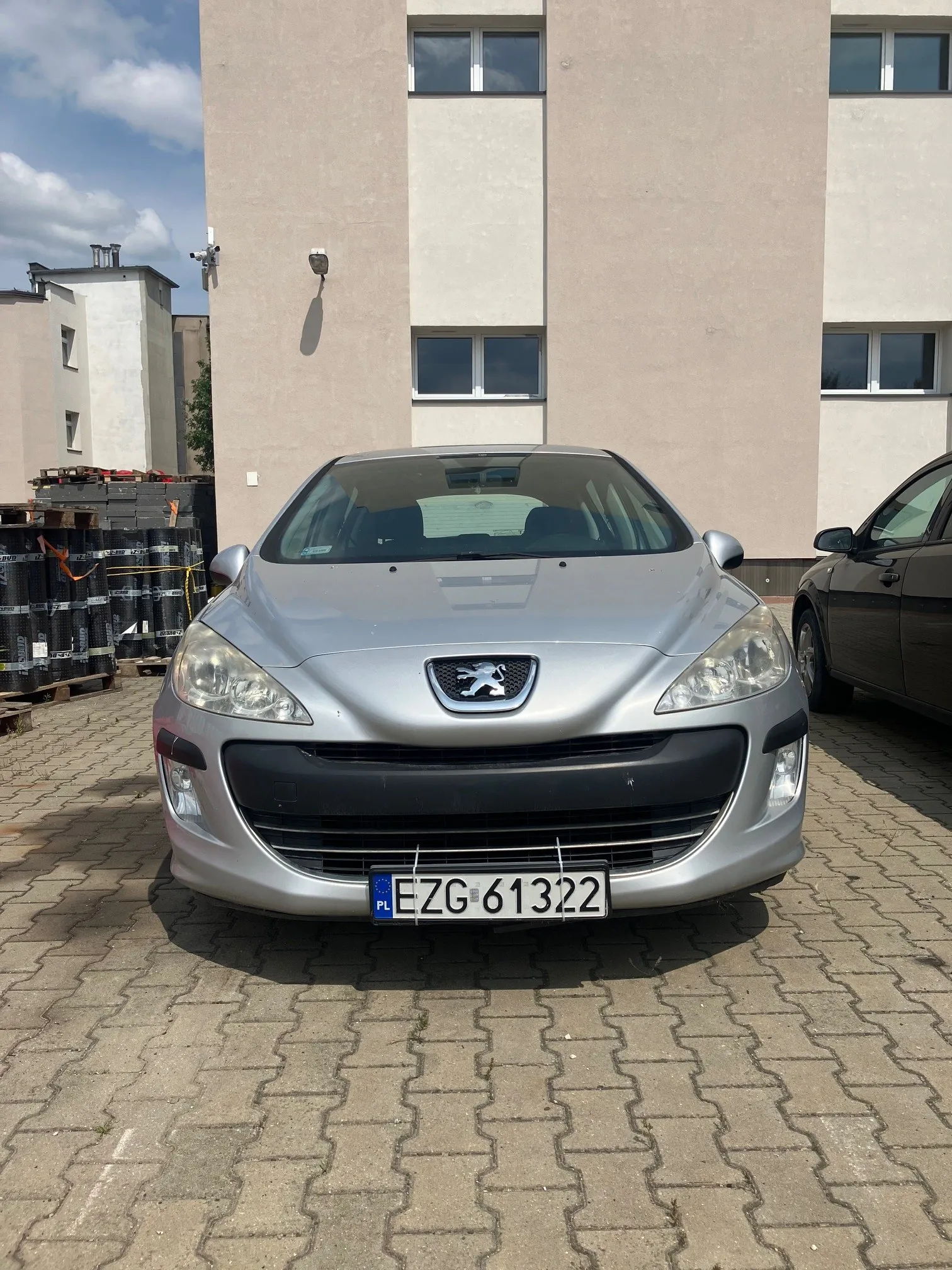 Srebrny samochód marki Peugeot zaparkowany przodem do widza na kostce brukowej przed jasnym budynkiem mieszkalnym. Na przodzie auta widoczna jest tablica rejestracyjna o numerze EZG 61322 z oznaczeniem PL. Po lewej stronie znajdują się czarne palety z materiałami budowlanymi, a po prawej fragment innego, ciemnego samochodu.