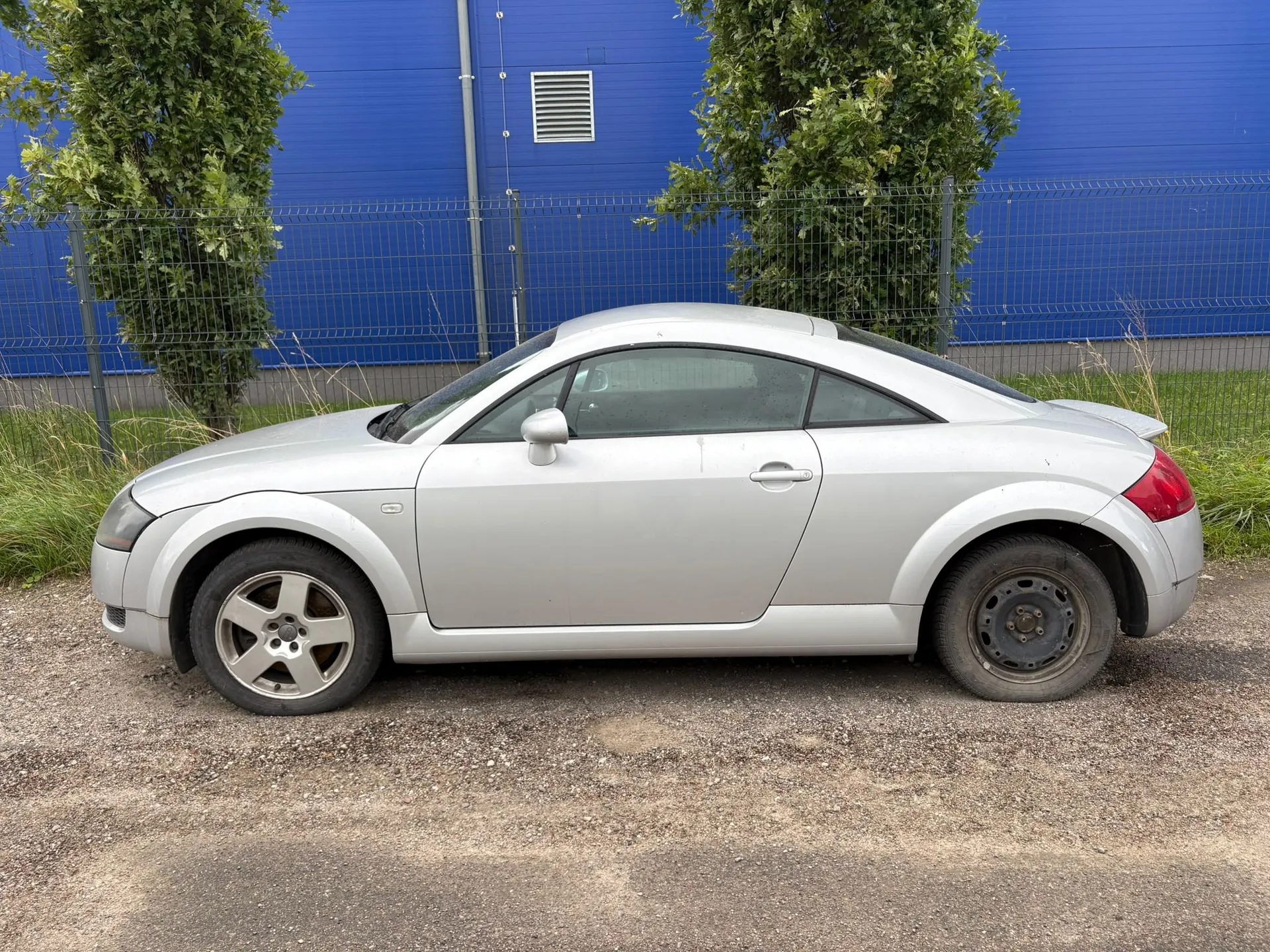 Srebrny samochód osobowy typu coupe zaparkowany bokiem na żwirowym podłożu. W tle znajduje się niebieska ściana budynku, metalowy płot oraz dwa zielone drzewa.