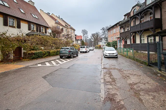 Zdjęcie przedstawiające wąską ulicę w zabudowie mieszkalnej. Po obu stronach drogi znajdują się wielorodzinne domy o zróżnicowanej architekturze i kolorystyce elewacji. Wzdłuż ulicy zaparkowane są samochody: srebrny SUV po lewej stronie, biały furgon oraz biały samochód osobowy po prawej. Nawierzchnia drogi jest asfaltowa, z widocznymi łatami i pęknięciami po prawej stronie. Po lewej stronie znajduje się przejście dla pieszych z białymi pasami. Niebo jest zachmurzone i szare.