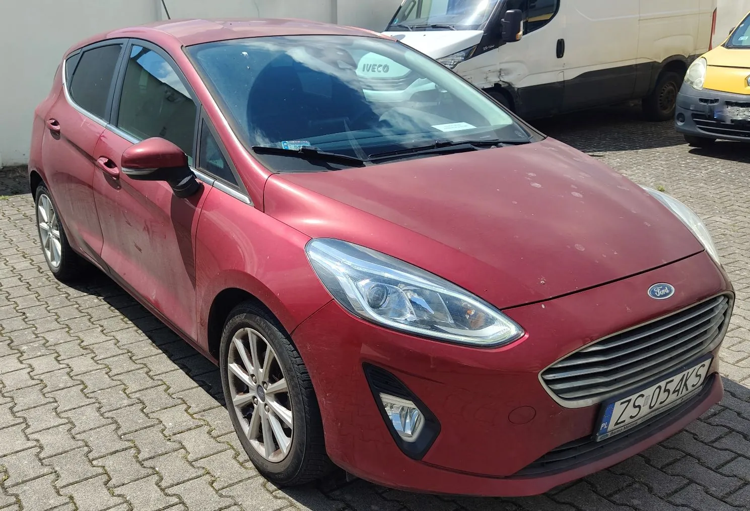 Czerwony samochód marki Ford zaparkowany na kostce brukowej. Pojazd jest widoczny z przodu i z boku. Na masce widoczne są drobne zanieczyszczenia. Samochód posiada polską tablicę rejestracyjną o numerze ZS 054KS. W tle widoczny jest biały samochód dostawczy marki Iveco oraz fragment żółtego pojazdu.